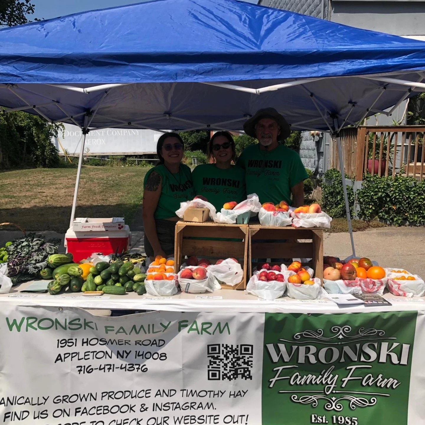 Three people standing behind a table of fresh produce at a farmers market stall under a blue canopy, with a banner displaying the farm's contact information and name, 'Wronski Family Farm.' The table has various fruits and vegetables, including peach