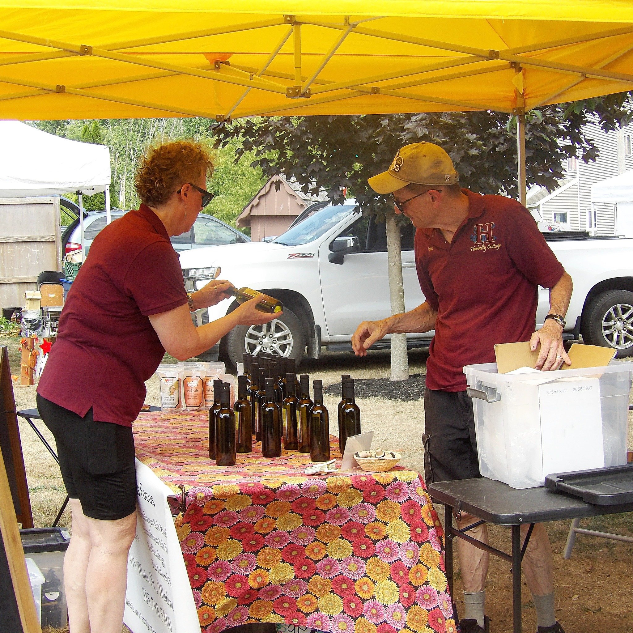 A woman and a man at an outdoor market stand with a yellow canopy, arranging bottles on a table covered with a colorful floral cloth. The woman is holding a device, possibly barcode scanner or calculator, and the man is reaching into a large transpar