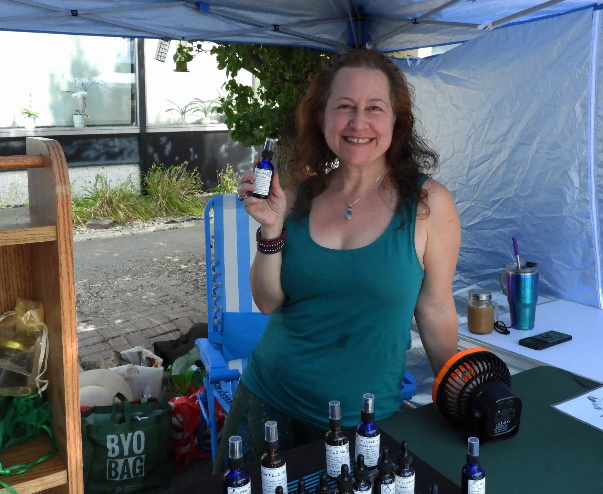 A smiling woman with light skin, curly reddish-brown hair, wearing a teal tank top, standing under a blue canopy tent. She holds a small bottle in her right hand. In front of her is a display of various small bottles with droppers, and a fan on the t