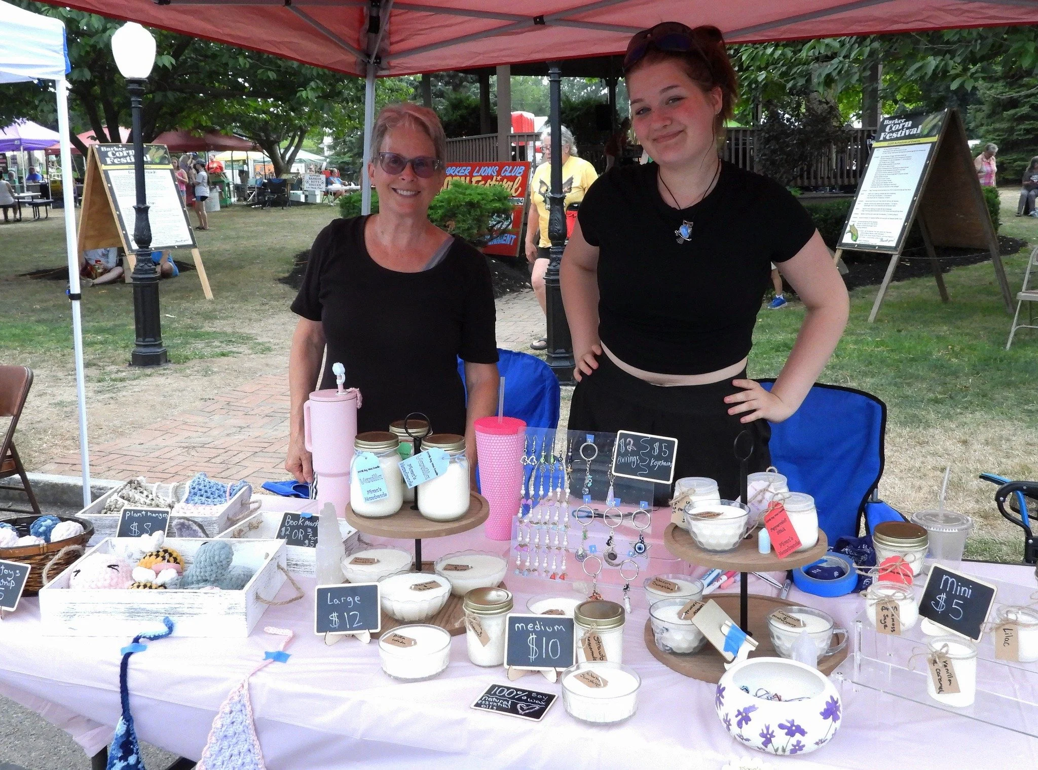 Two women standing behind a table at an outdoor craft fair, displaying handmade jewelry and candles under a red canopy. One woman is wearing sunglasses and the other has her hands on her hips. The table has various jars, candles, and jewelry with sma