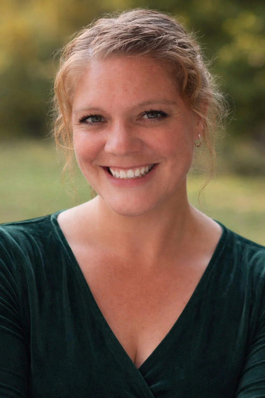 A smiling woman with light red hair in a loose updo, wearing small hoop earrings and a dark green top, outdoors with a blurred green and yellow background.