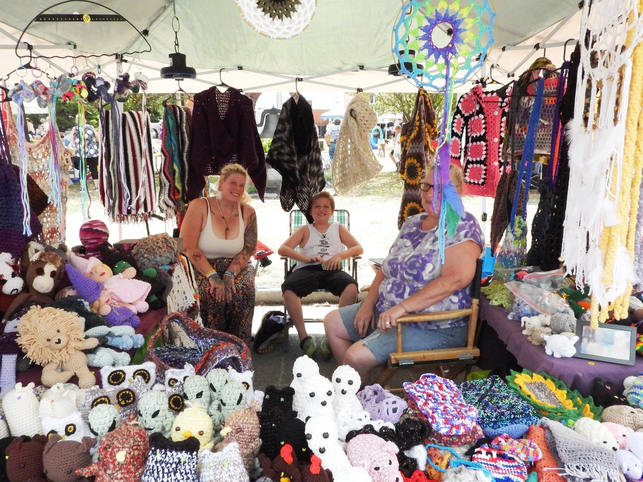 Three women sitting at a craft fair booth with handmade knitted crocheted stuffed animals, hats, and clothing on display.