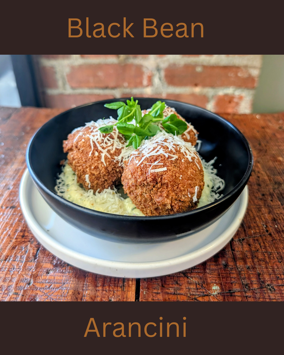 A black bowl of arancini topped with shredded cheese and garnished with fresh herbs, served on a white plate on a rustic wooden table.