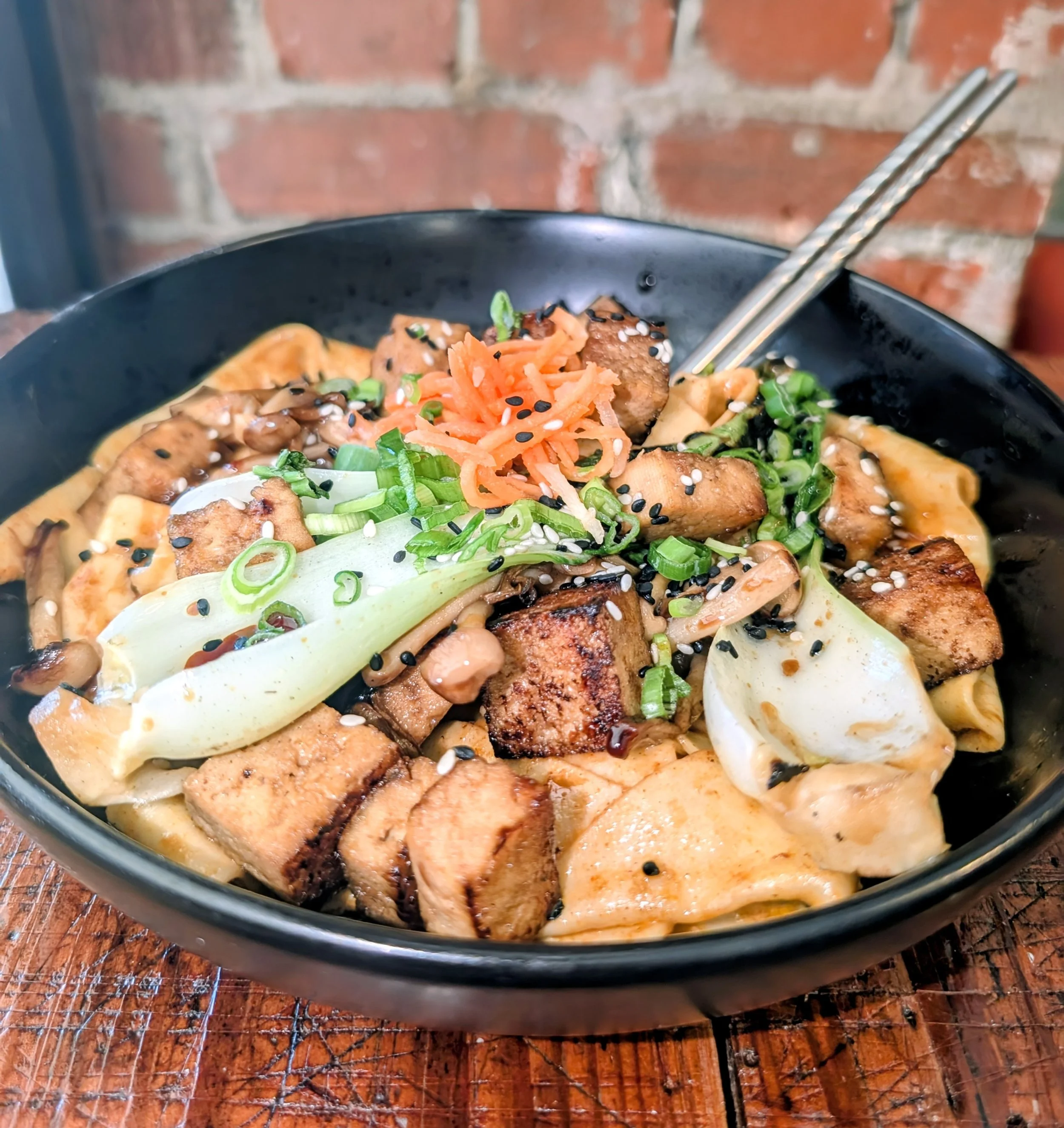 A black bowl of Asian noodle soup topped with grilled chicken, shredded carrots, chopped green onions, black sesame seeds, and white sesame seeds, with chopsticks resting on the bowl's edge against a brick wall background.