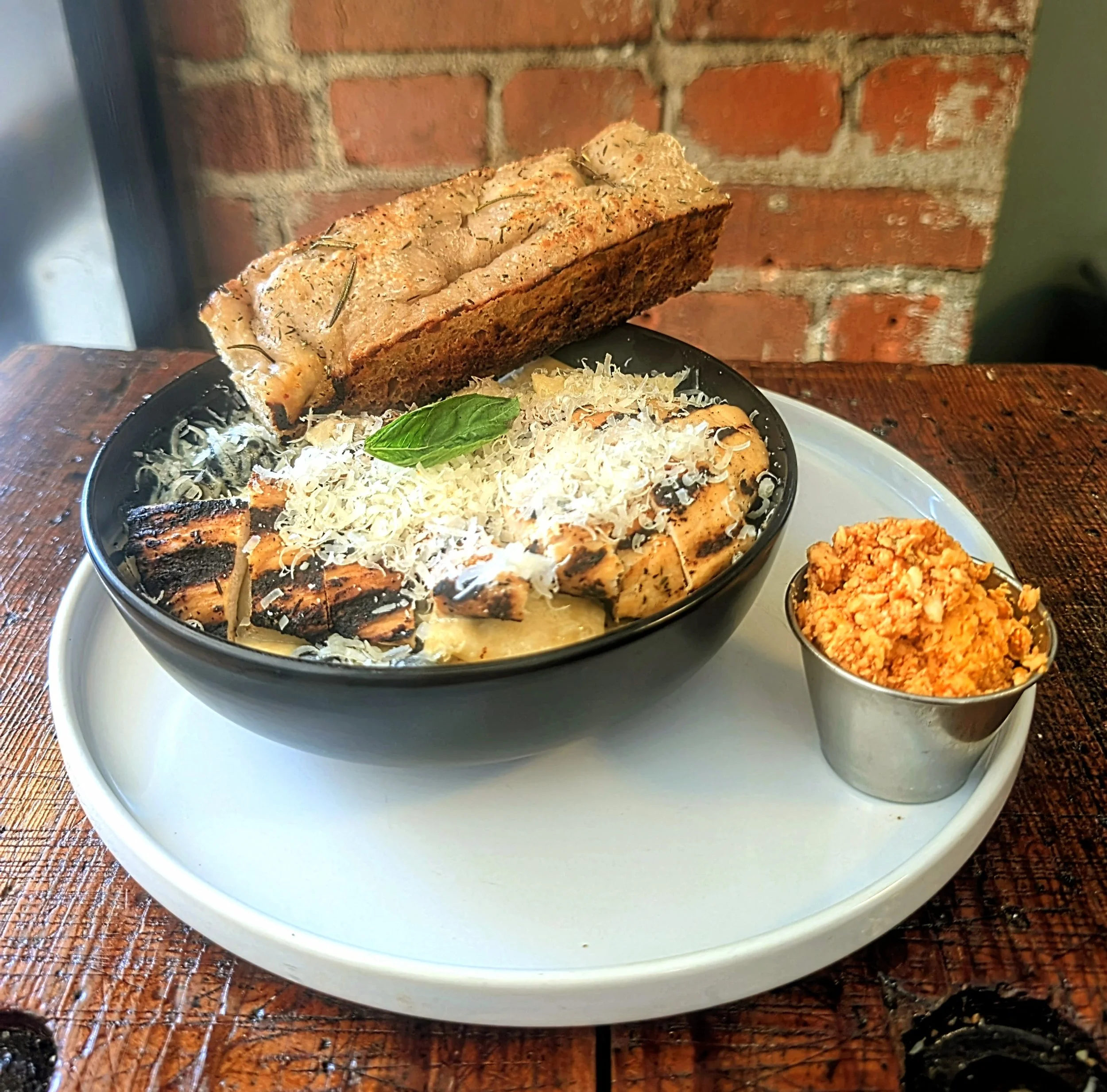 A plate of grilled chicken breasts topped with shredded cheese and a basil leaf, accompanied by roasted vegetables, with a loaf of baked bread topped with herbs, set against a brick wall background.