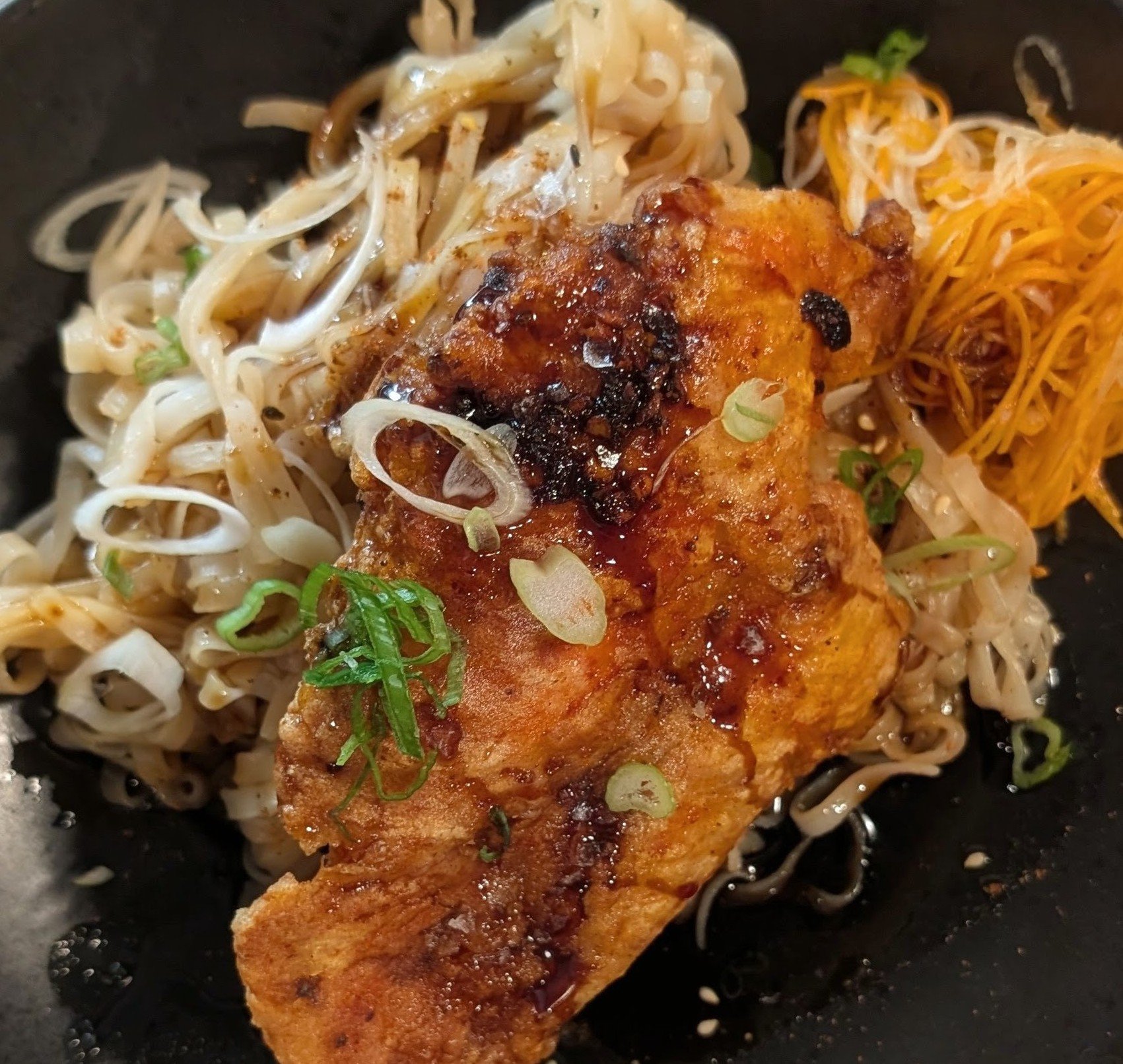 Fried fish fillet topped with sauce and green onions, served on a bed of noodles or shredded vegetables, in a dark bowl.