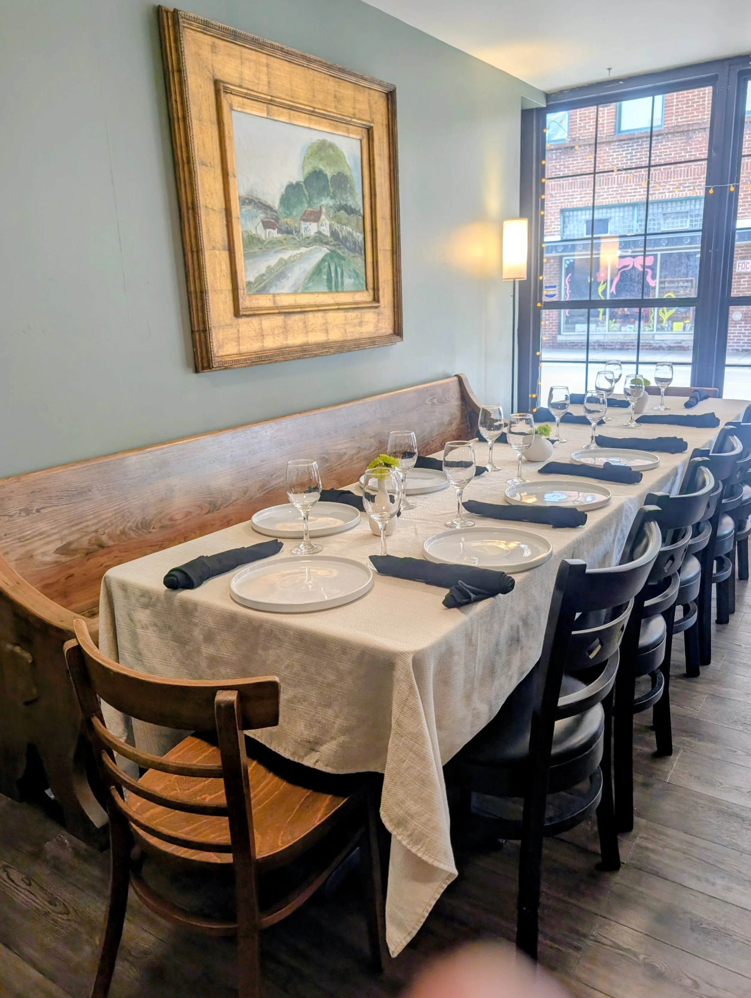 A long dining table set for a meal with wine glasses, plates, black napkins, and small decorative centerpieces, in a restaurant with large windows and artwork on the walls.