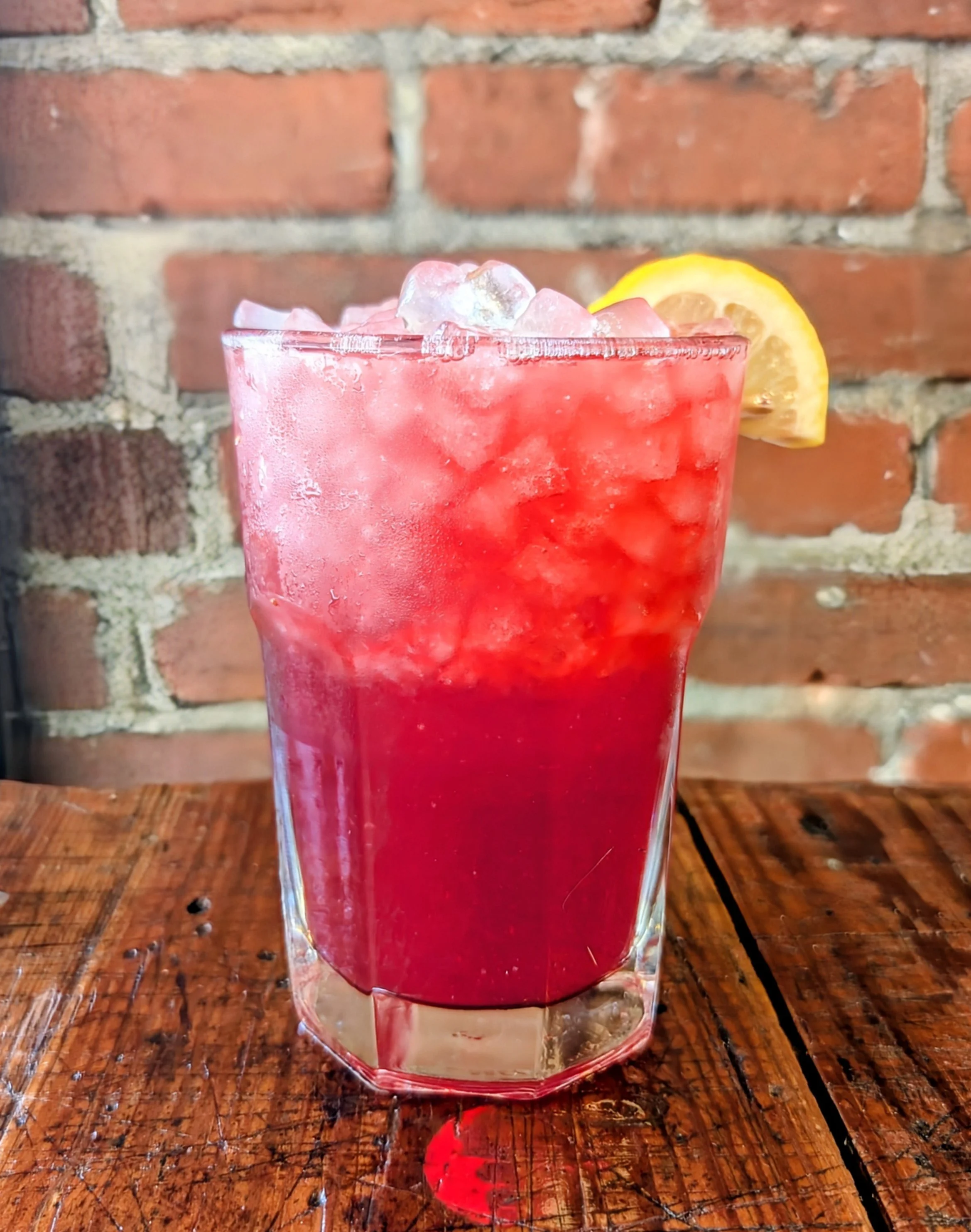 A glass of red frozen drink with ice cubes and a lemon wedge on the rim, on a wooden table in front of a brick wall.