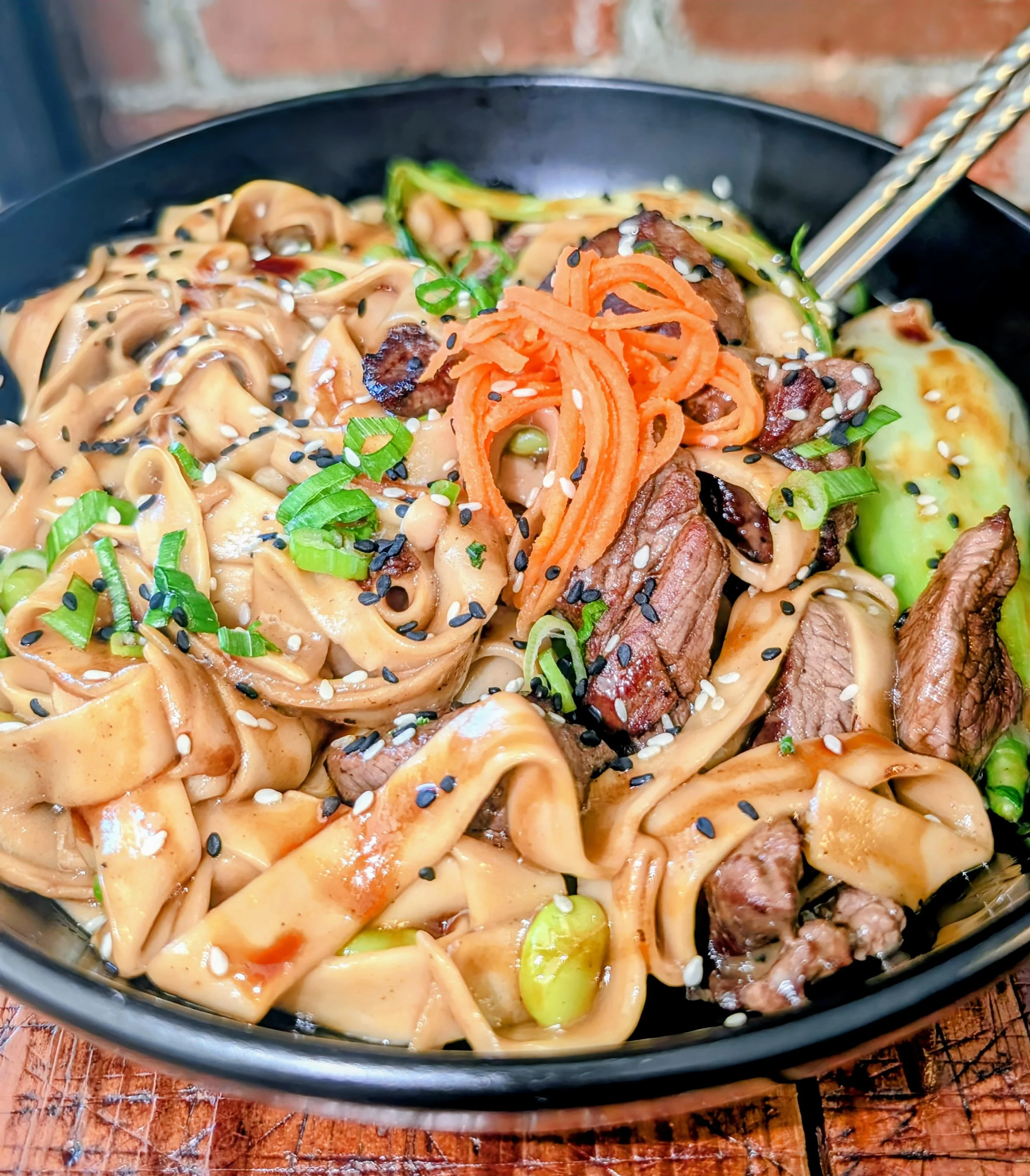 A black bowl of Asian-style noodles with sliced beef, shredded carrots, green onions, cucumbers, sesame seeds, and a light sauce, set on a wooden surface.
