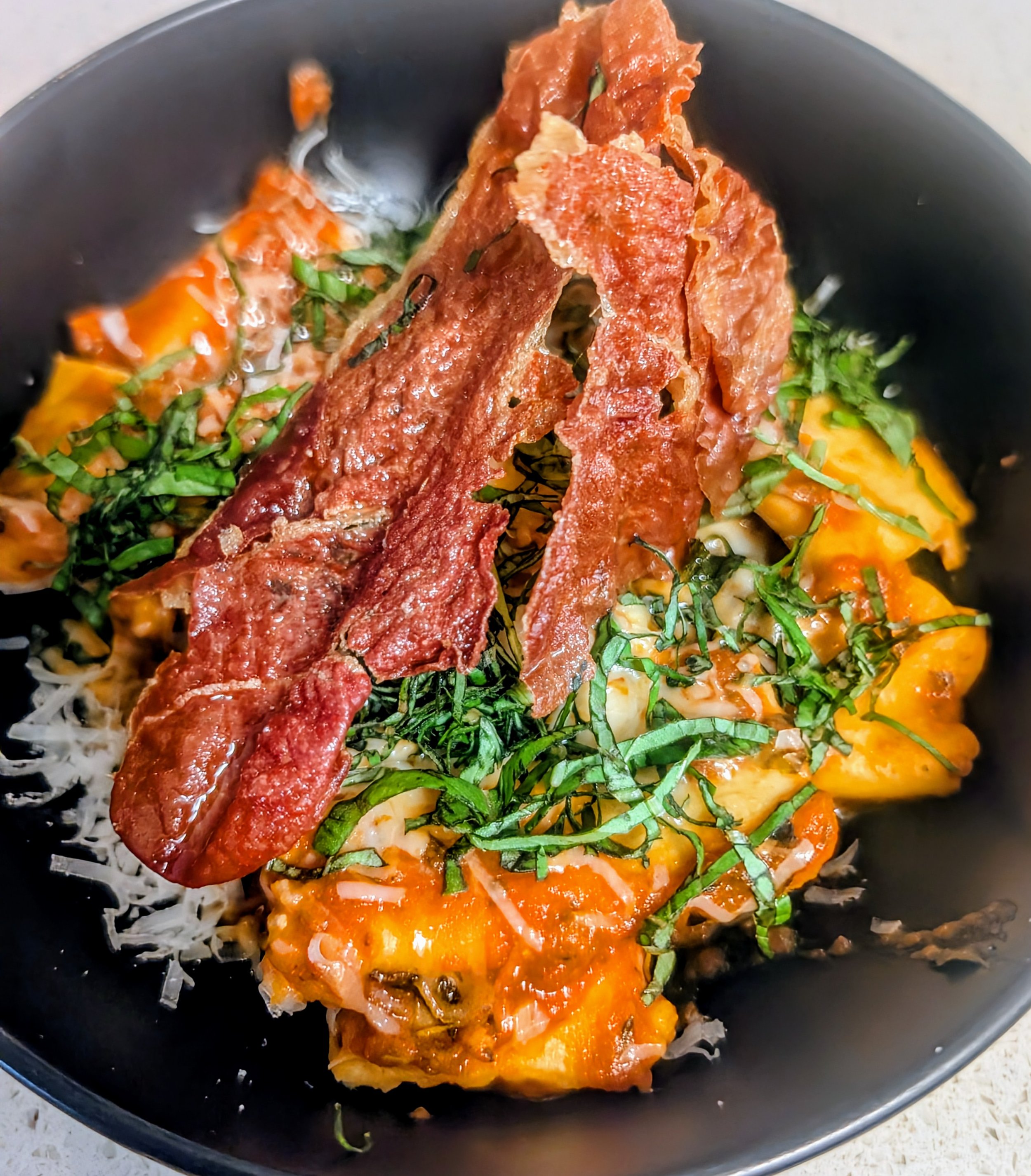 A black skillet containing cooked eggs, avocado slices, chopped green herbs, and crispy bacon strips.