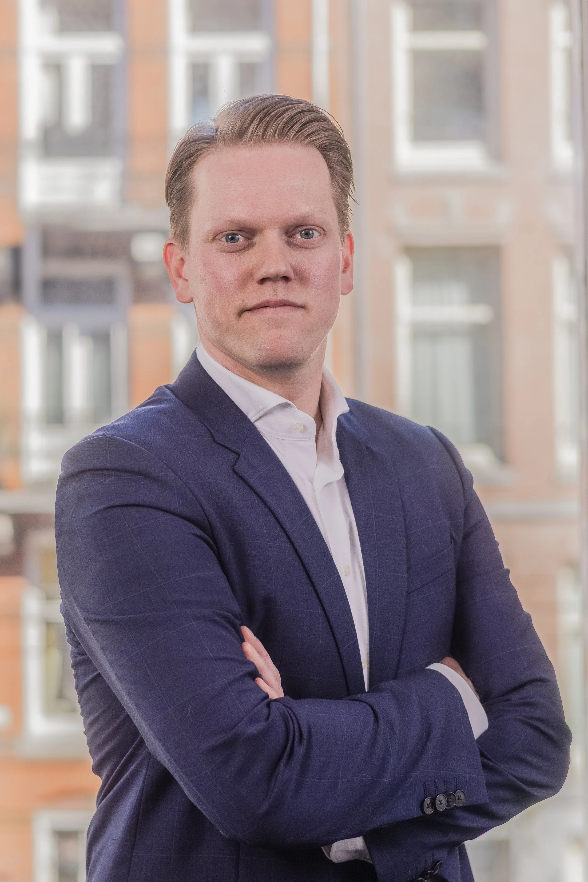 Een man in zakelijke kleding met gekruiste armen, staand voor een raam met een stedelijk uitzicht.