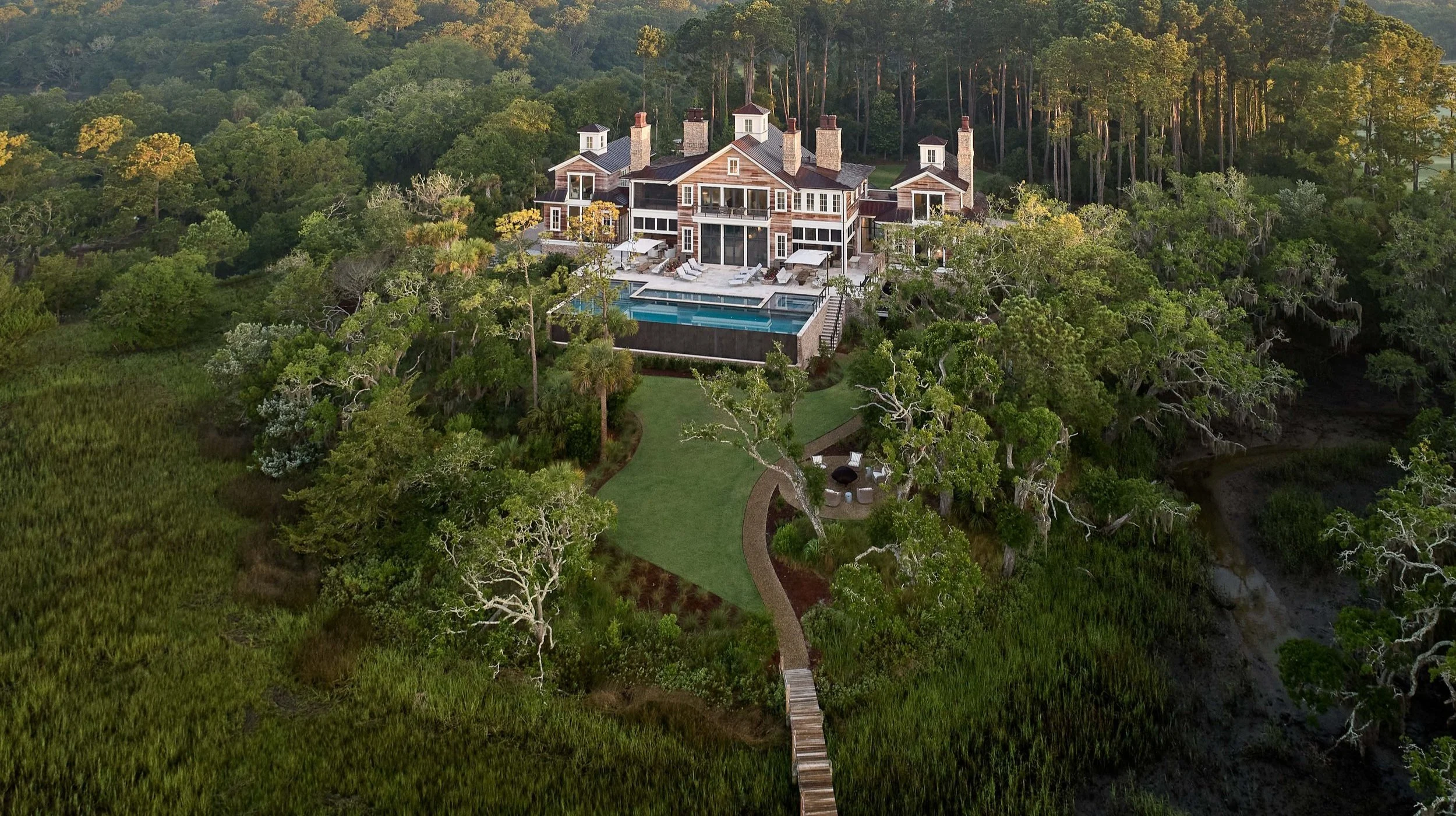 Large house with multiple chimneys, balconies, and a pool, surrounded by dense trees and greenery.