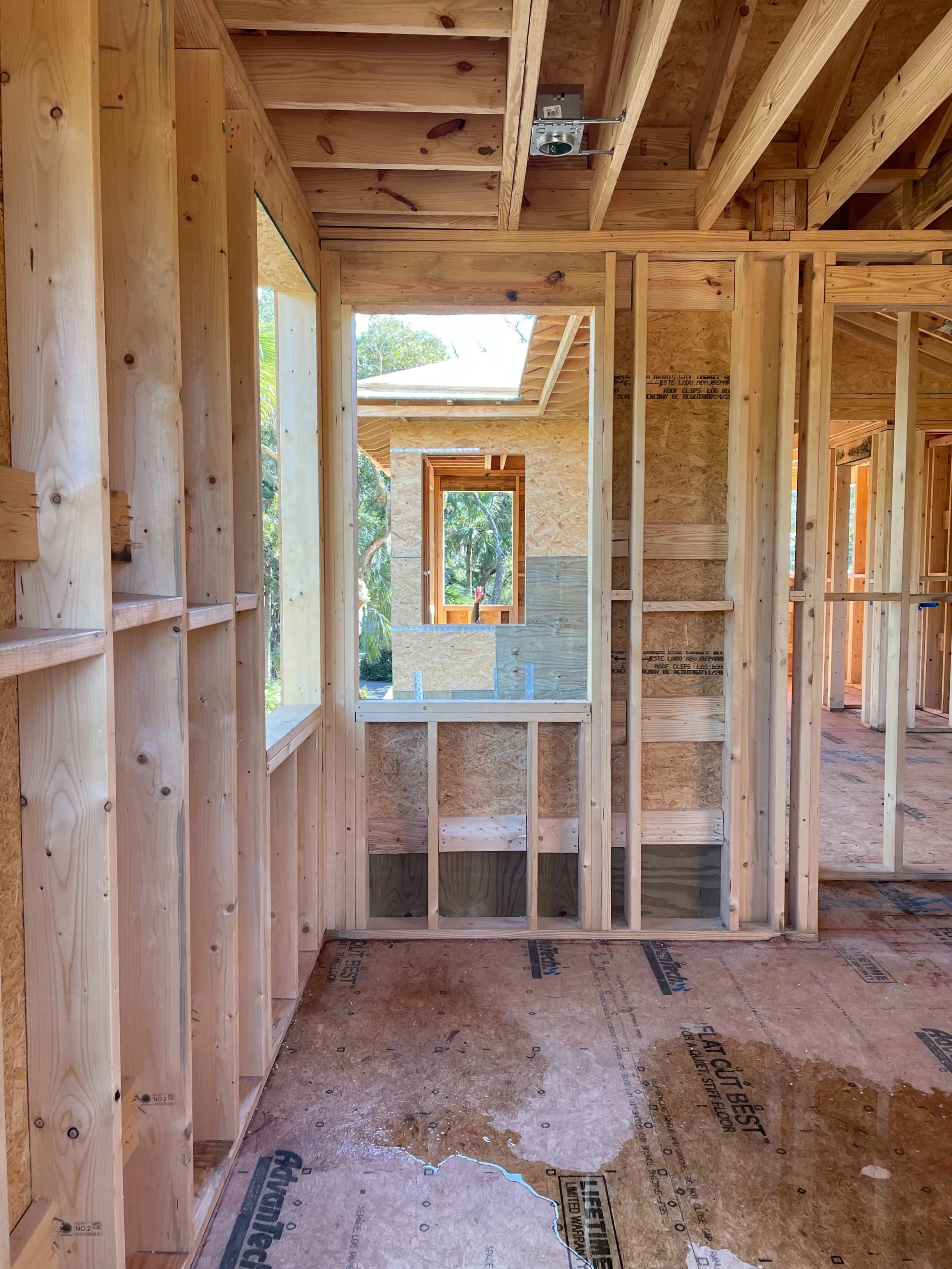 Interior view of a house under construction showing wooden framing, window openings, and unfinished flooring.