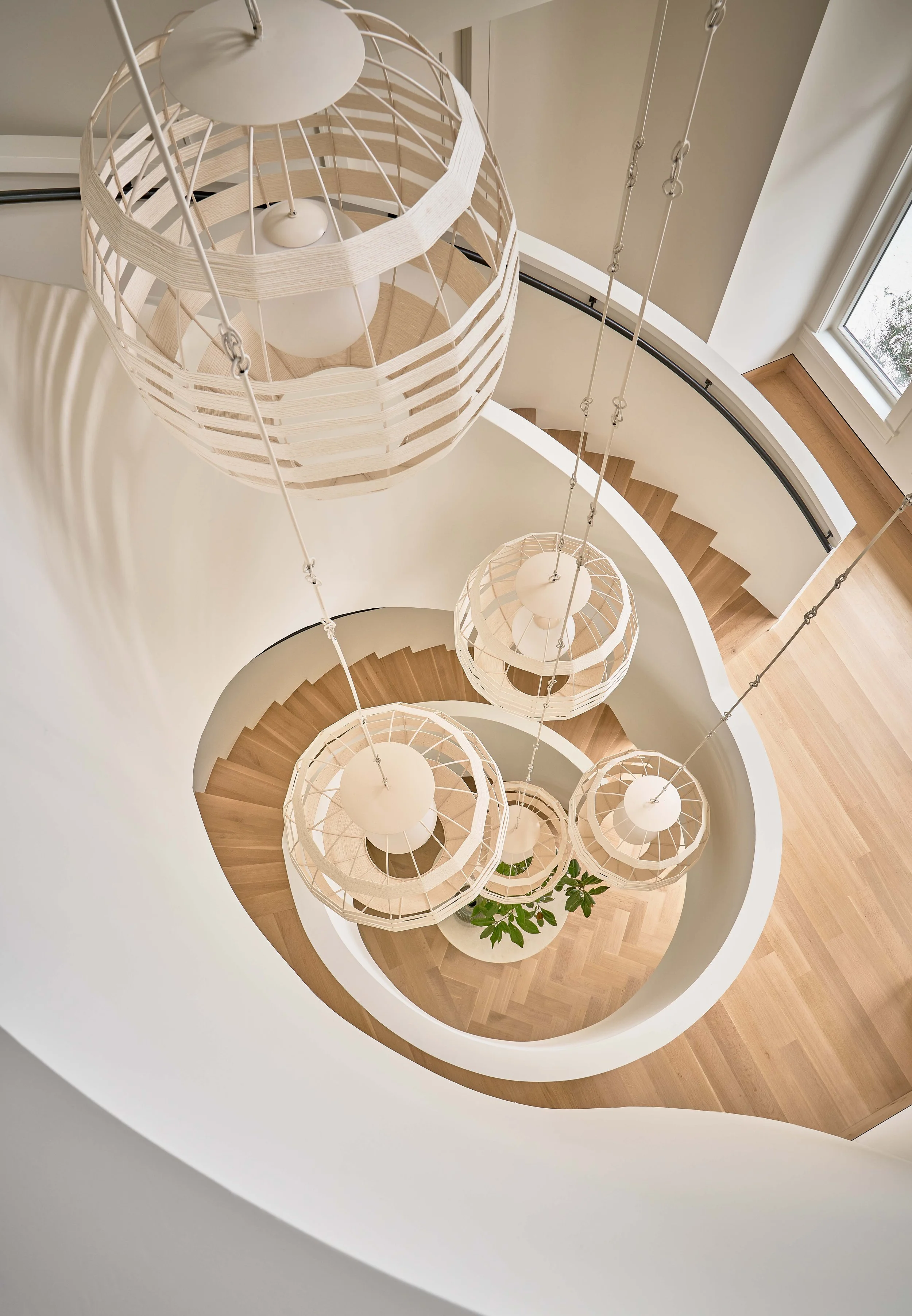 Looking down at a spiral staircase with five hanging pendant lights and a potted plant at the bottom near a window.