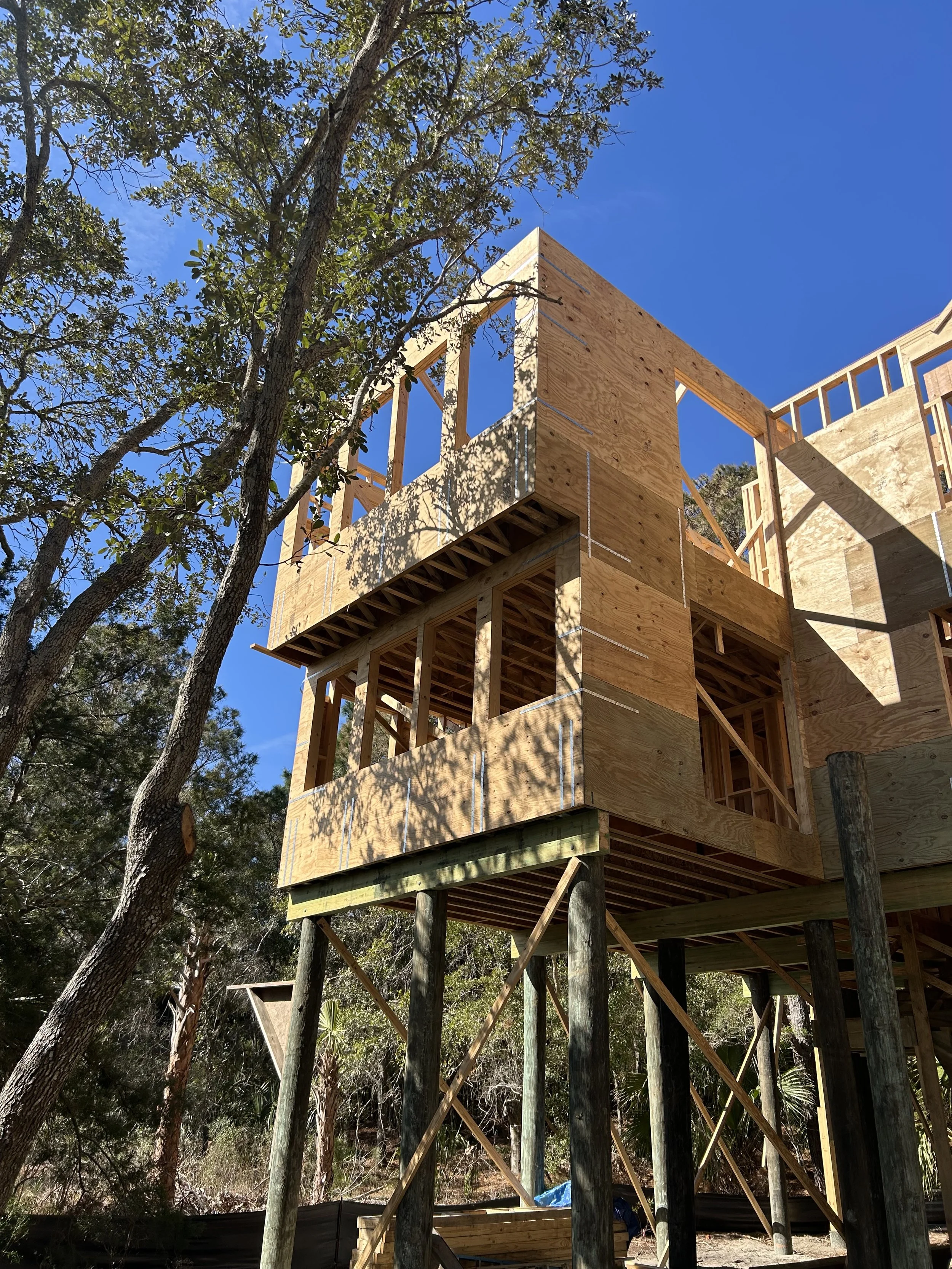 Under construction wooden house on stilts with framing and blue sky visible.