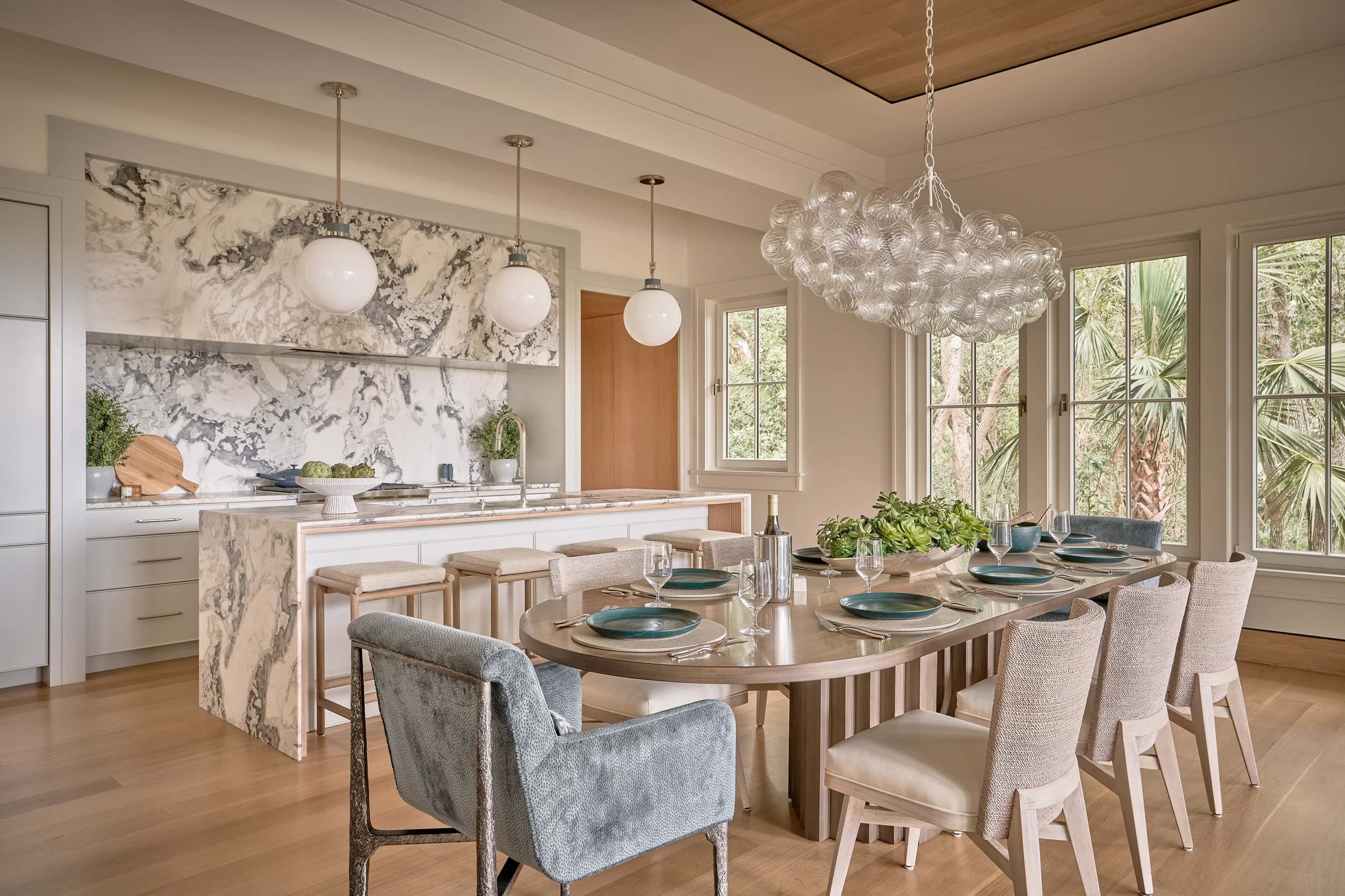 Modern dining area with a large wooden table set with black plates, wine glasses, and an arrangement of greenery in a white oval bowl. Multiple beige and gray upholstered chairs surround the table. The background features a spacious kitchen with white cabinetry, a marble backsplash, a marble countertop island, and hanging white globe pendant lights. Large windows reveal lush greenery outside.