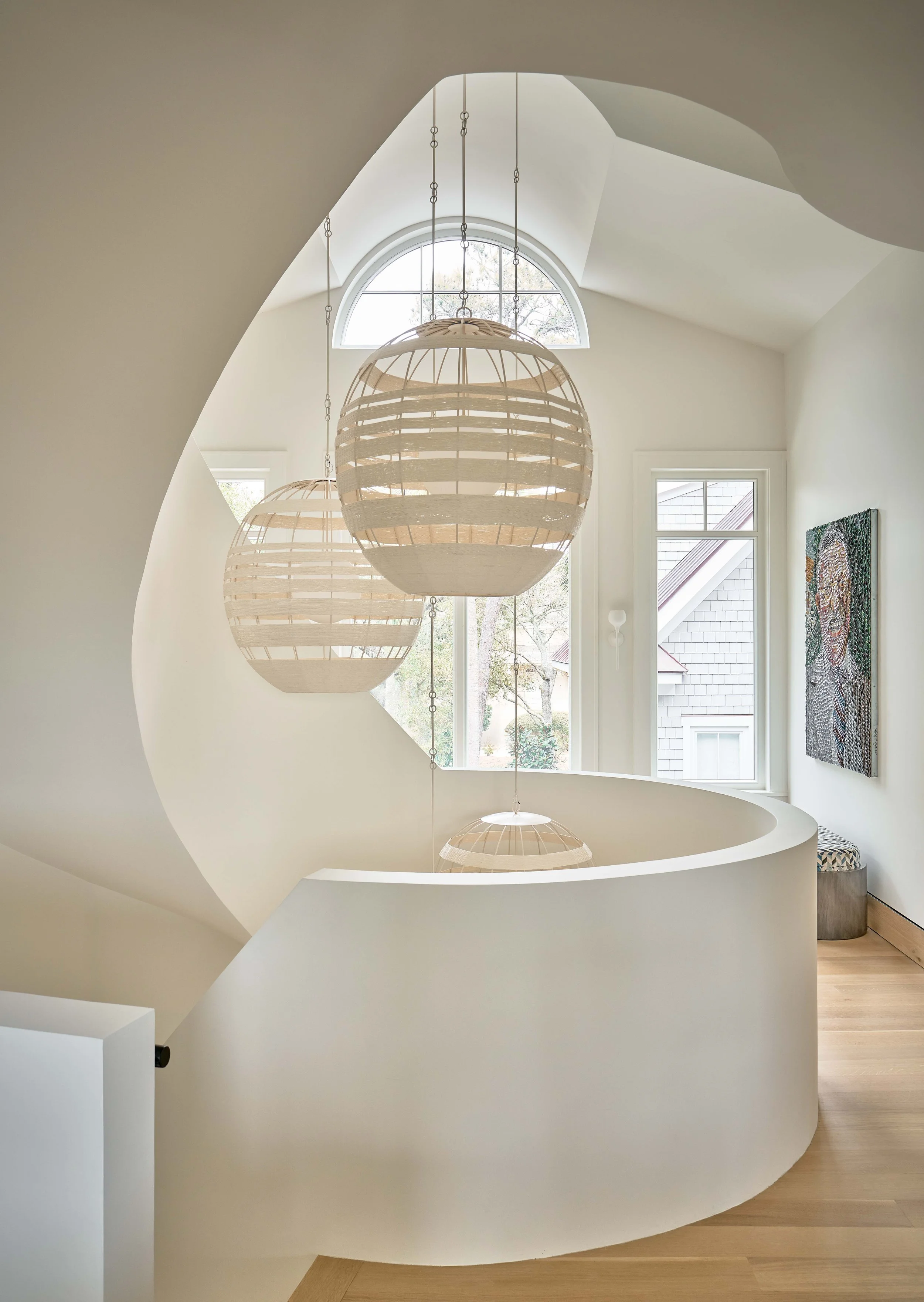 Interior view of a modern staircase with three hanging spherical pendant lights made of wooden slats, large windows allowing natural light, and a piece of colorful artwork on the wall.
