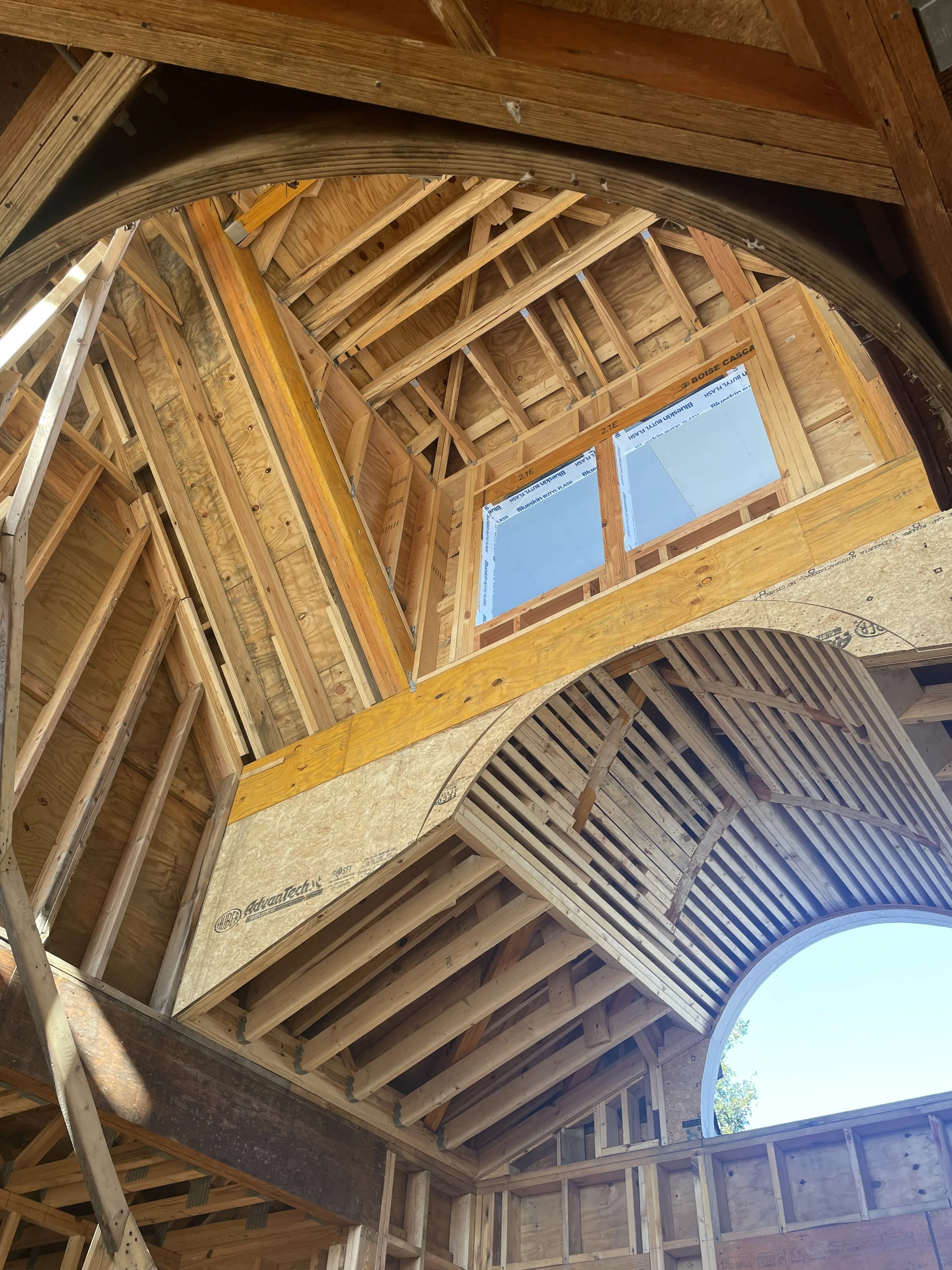 Interior view of a building under construction with exposed wooden framing, a curved wall opening, and a large window opening.