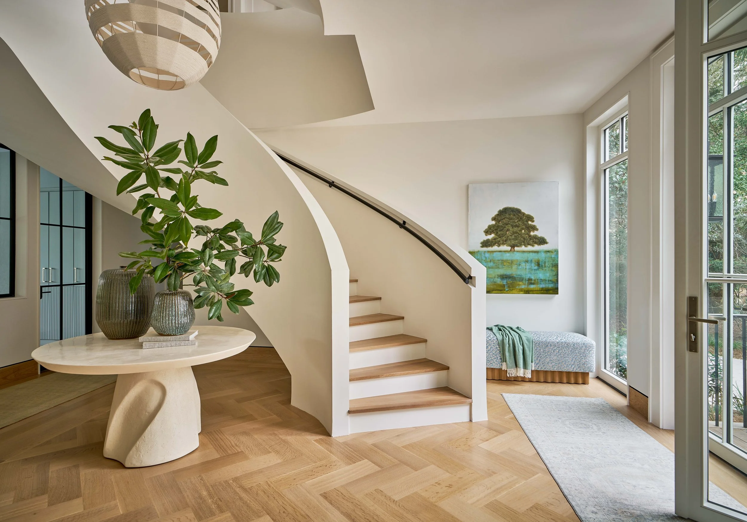 The interior of a modern house with a curved staircase, a round cream-colored table with vases and green plants, large windows, and a painting of a tree on the wall.