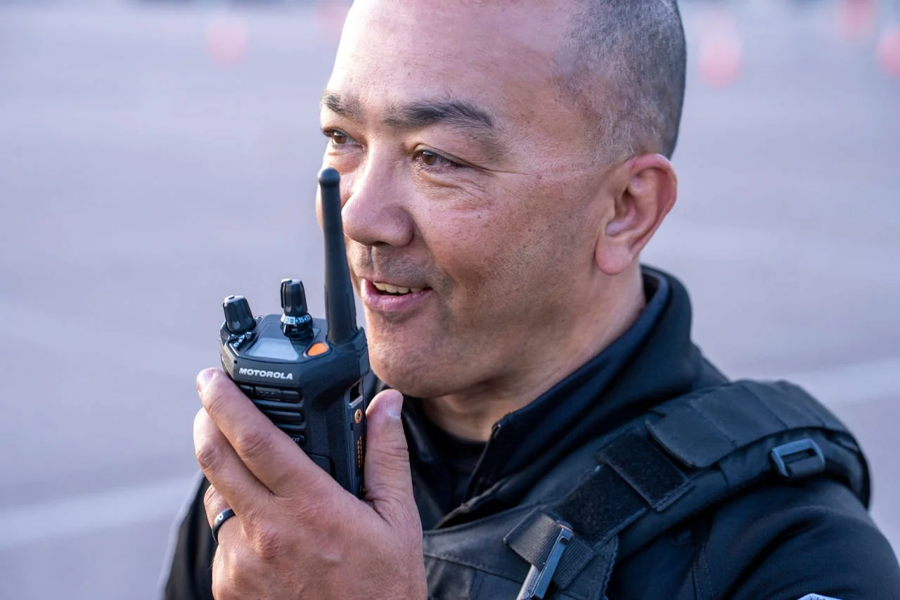 Close-up of a man holding a Motorola walkie-talkie to his mouth, speaking, with a blurred outdoor background.