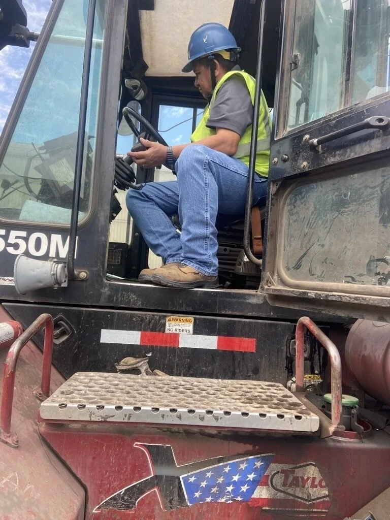 Construction worker sitting in a scissor lift, looking at a device in his hands, wearing a hard hat and safety vest.