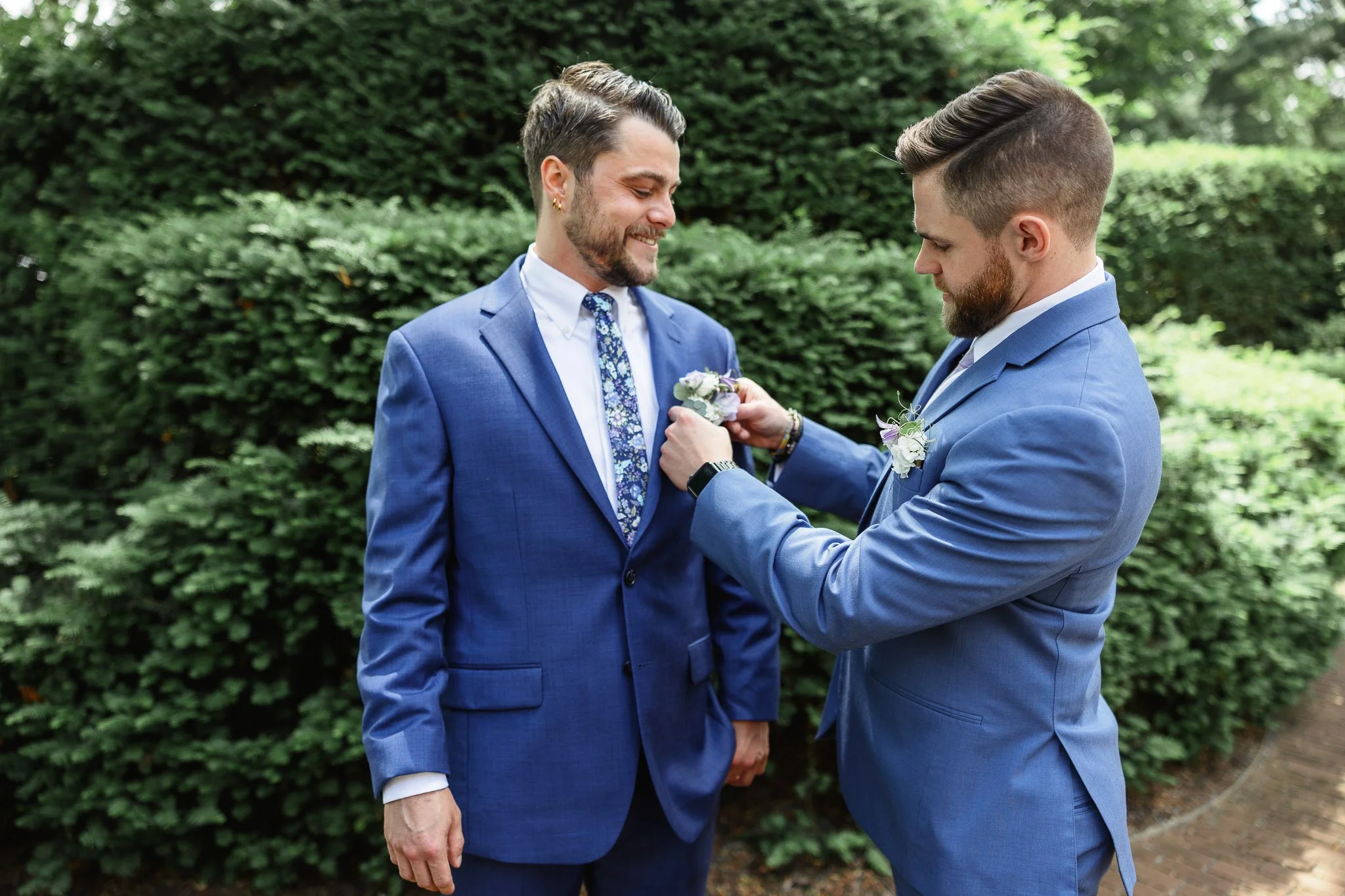 a wedding photo of a groomsman helps the groom adjust his boutonniere  