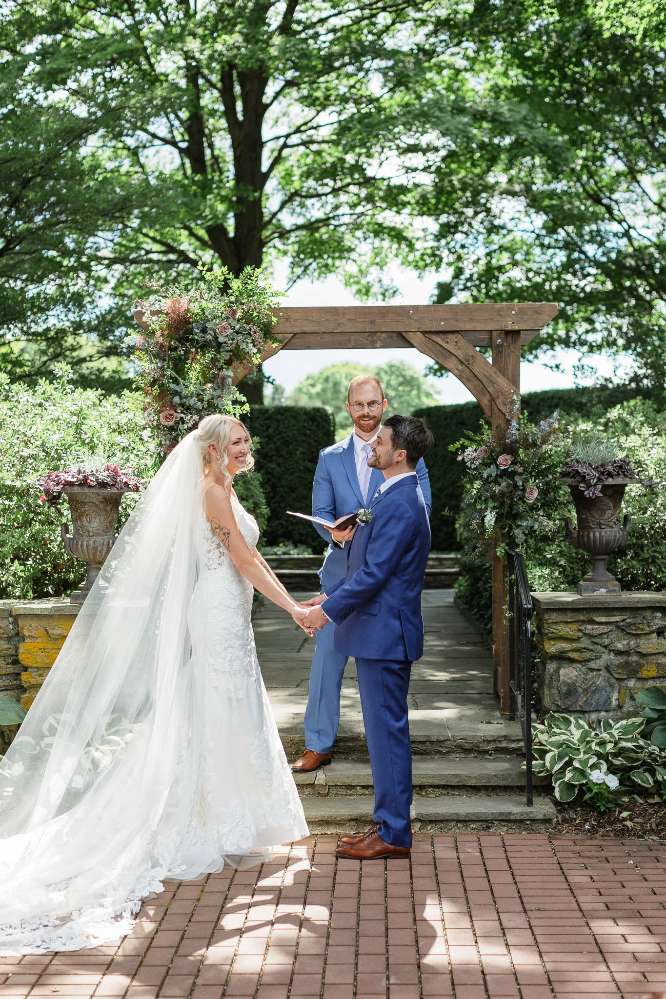 A bride in a white lacy gown and a groom in a blue suit exchange vows at The Gardens at Drumore Estate