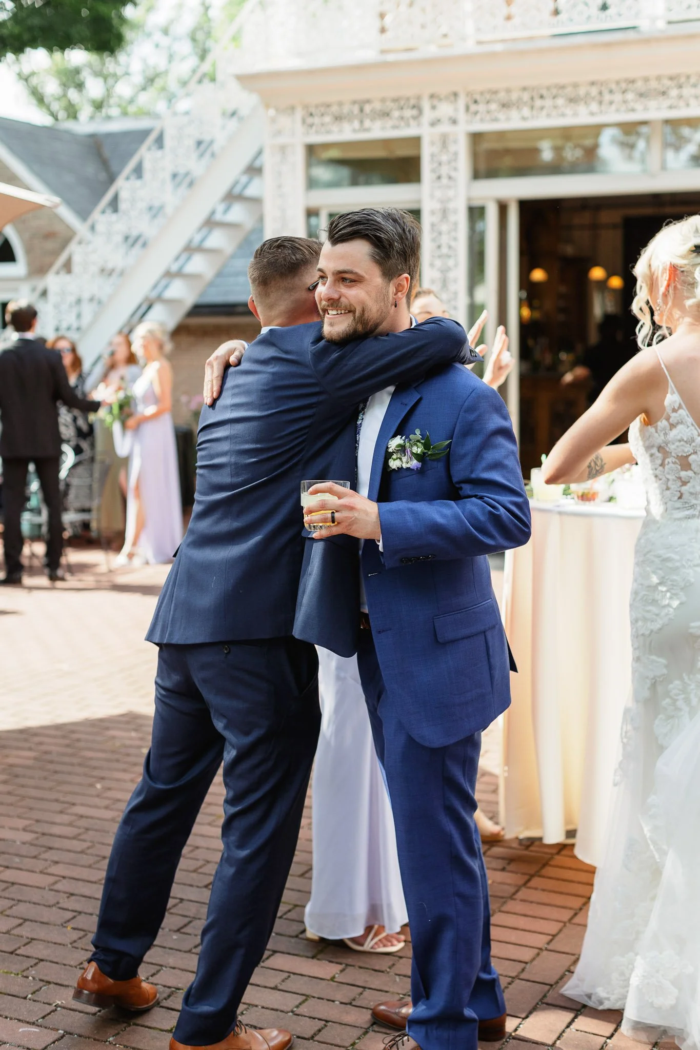 A wedding photographer captures the groom in his blue suit sharing a joyful embrace with a guest outside the venue.