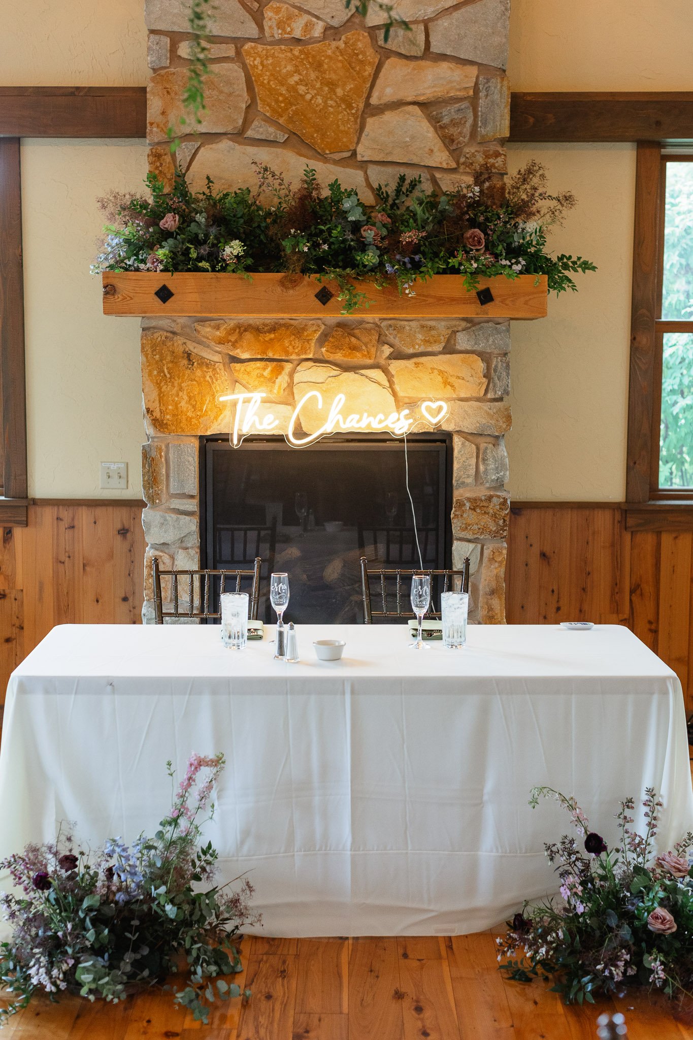 A cozy wedding photo of a grand stone fireplace at the venue, decorated with a neon sign and seasonal greenery to add warmth to the evening.