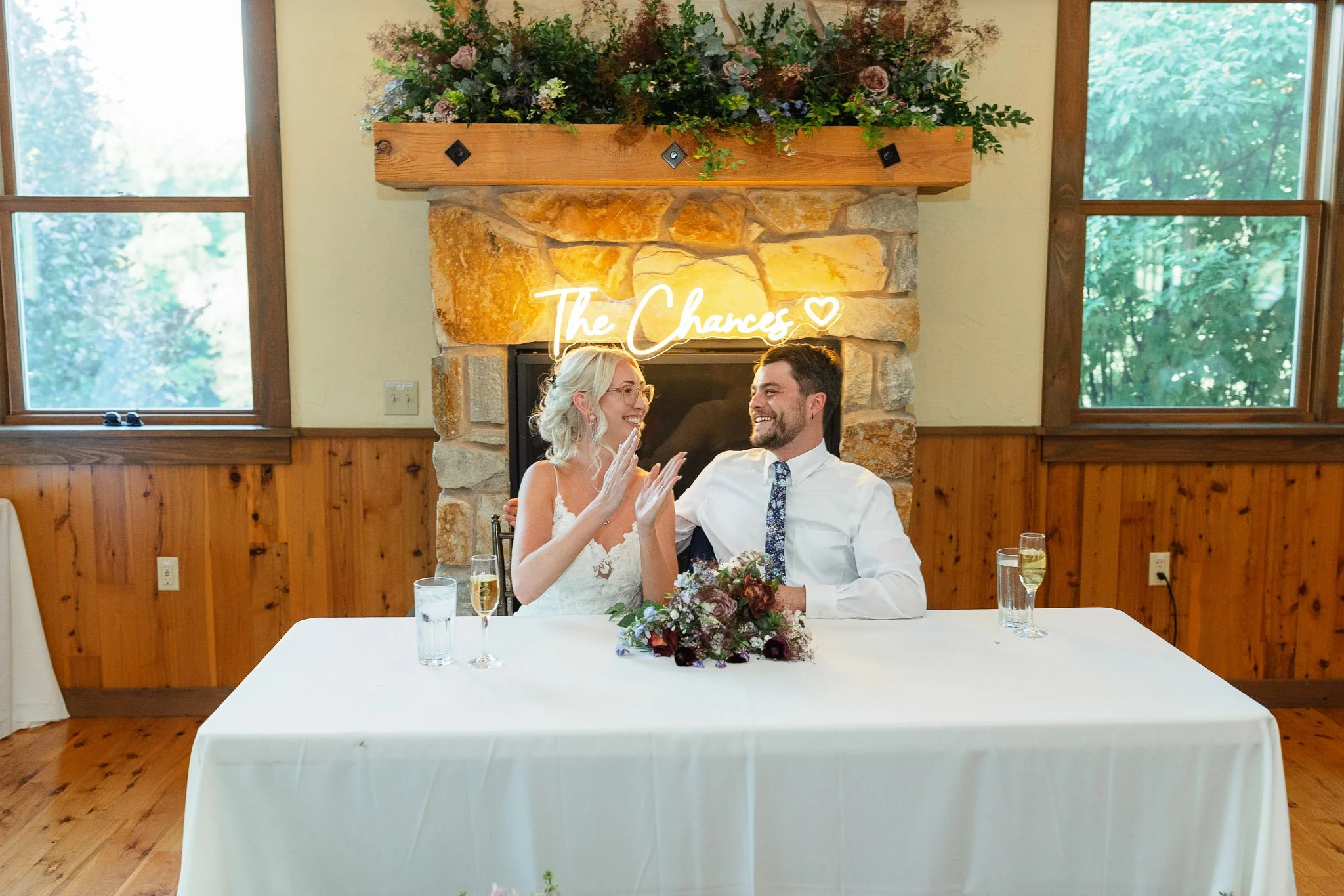 The groom in his blue suit looking at the best man while the bride in her white lace dress smiles during the reception toasts.
