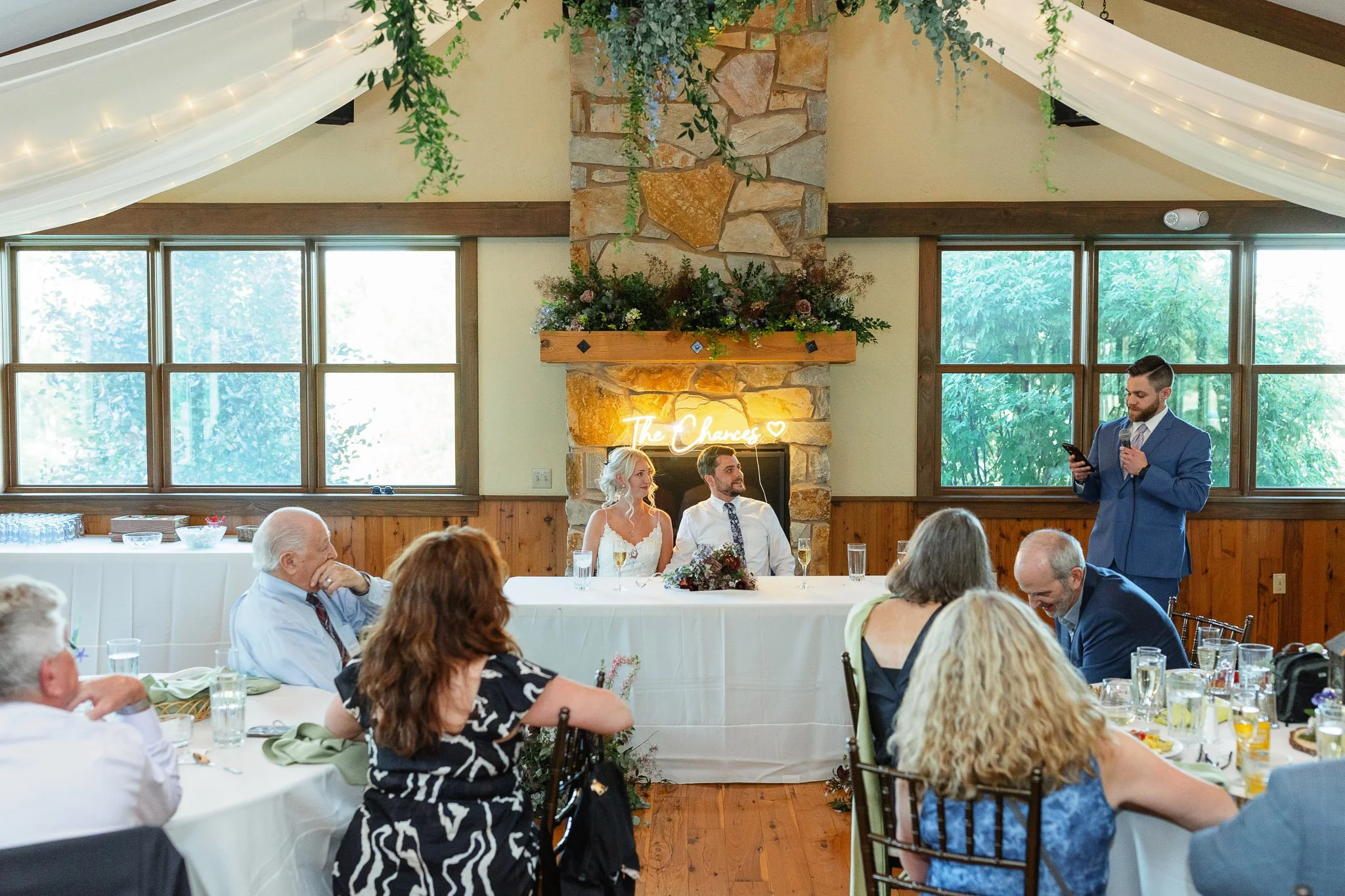 A wedding photo capturing the best man standing with a microphone while the bride and groom share a private smile at the head table.