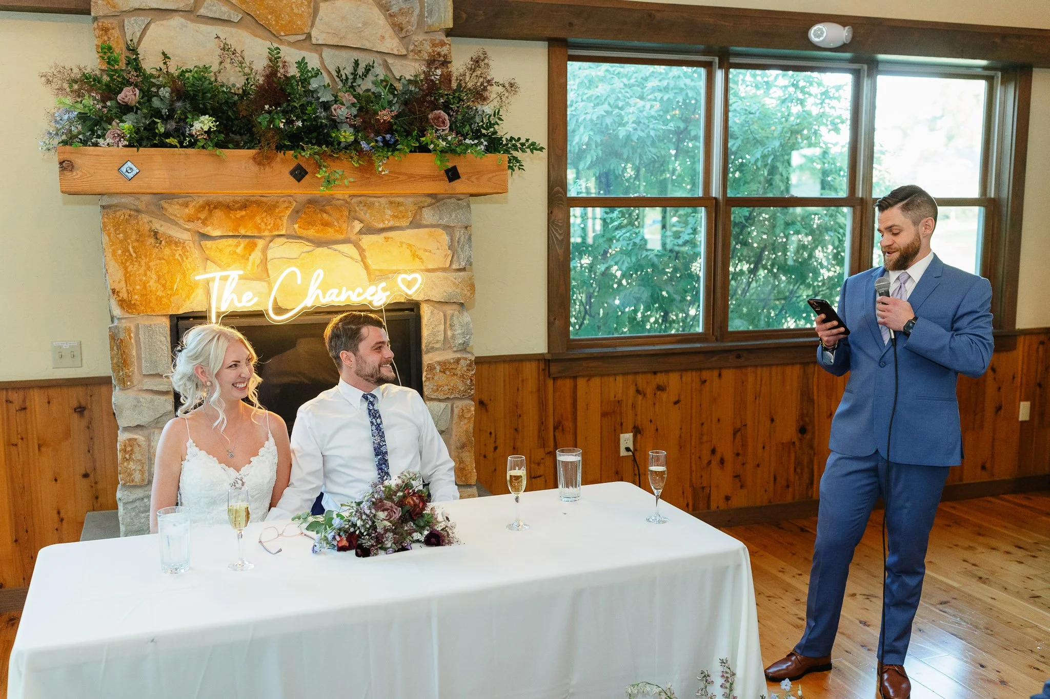 A professional wedding photographer's shot of the best man raising a glass toward the groom in his blue suit during a heartfelt speech.