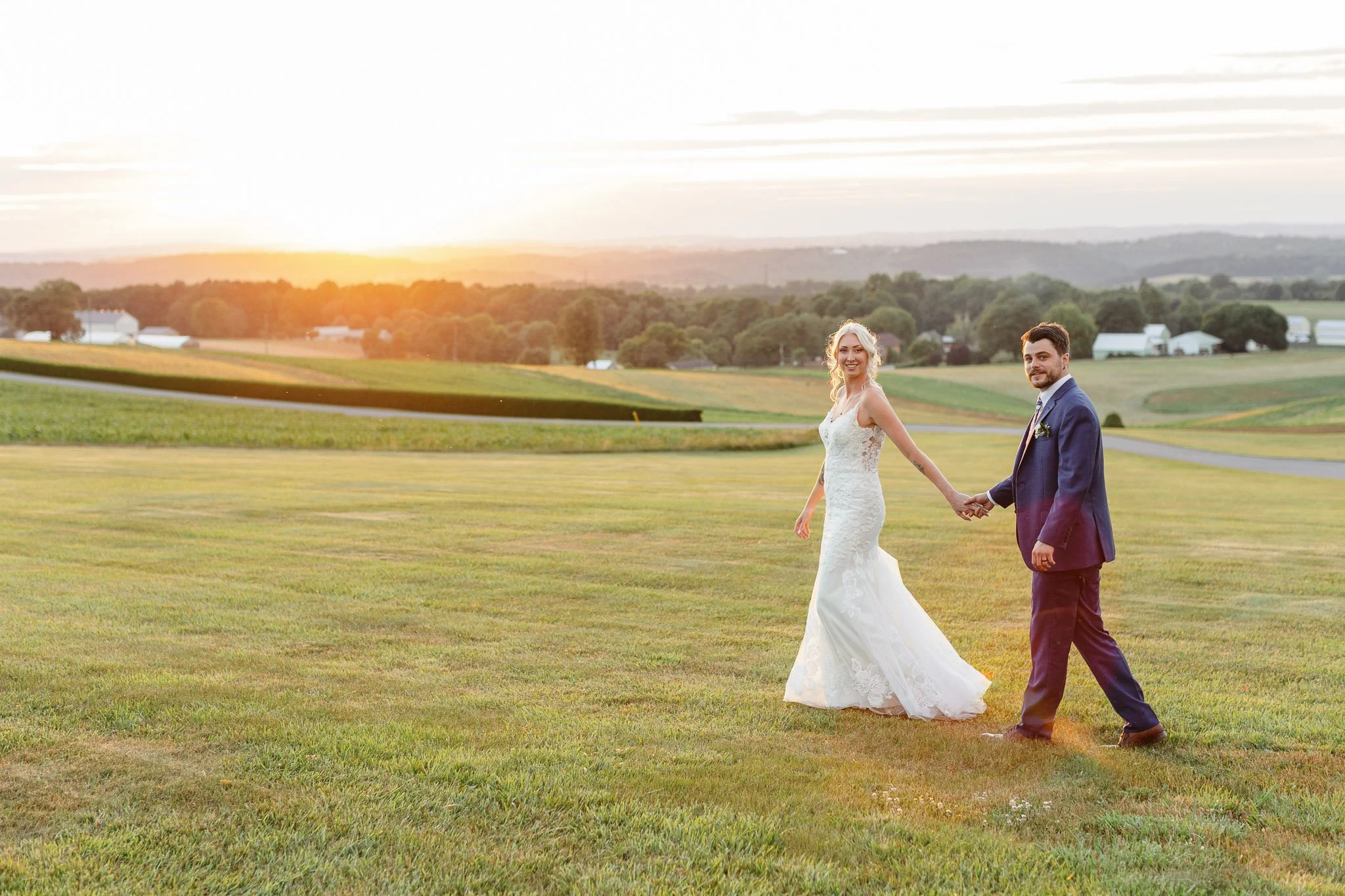 A wide-angle wedding photo of the bride and groom walking hand-in-hand across the Drumore Estate lawn at dusk.