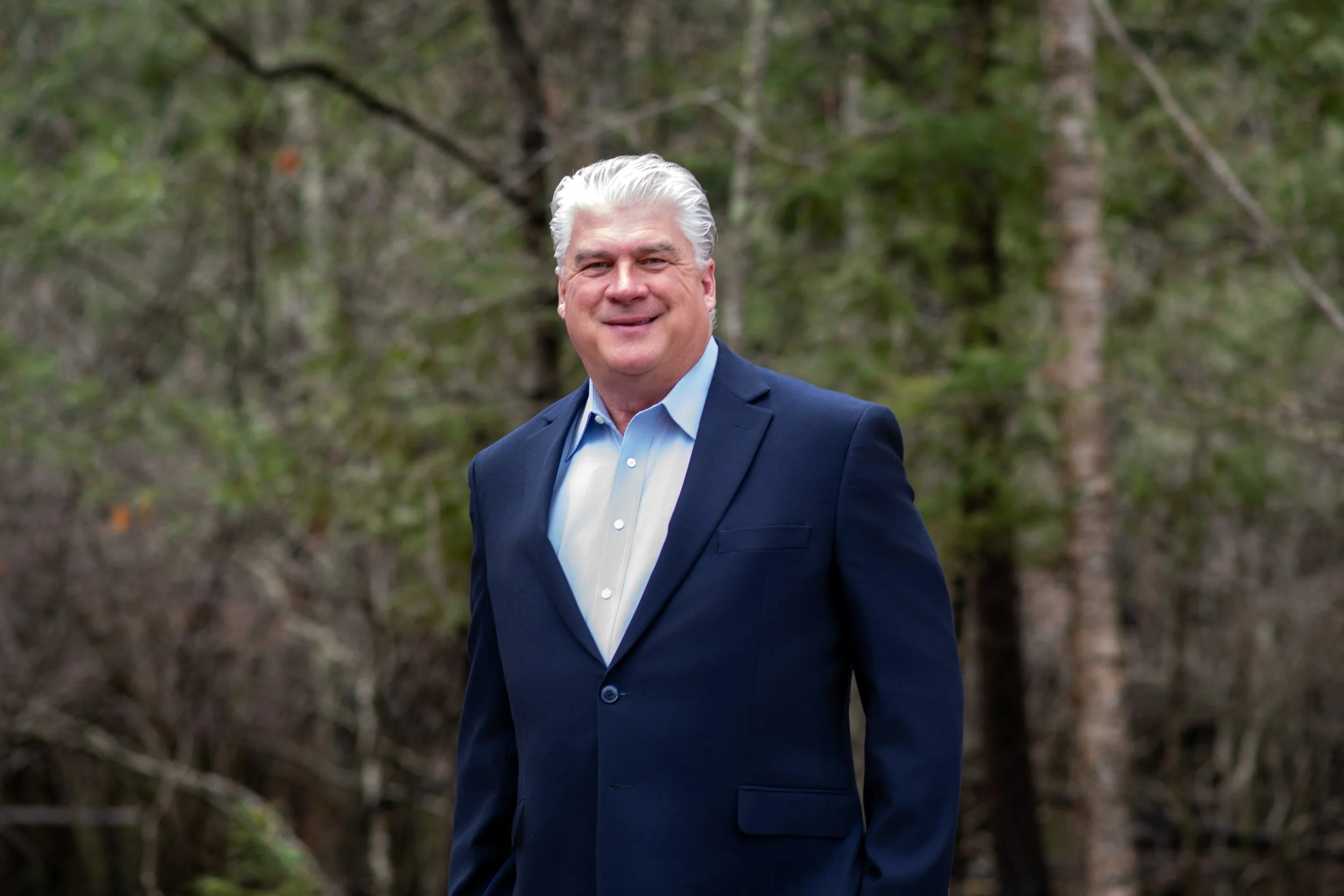 A smiling man with white hair in a navy blue suit and light blue shirt standing outdoors in front of a blurred forest background.