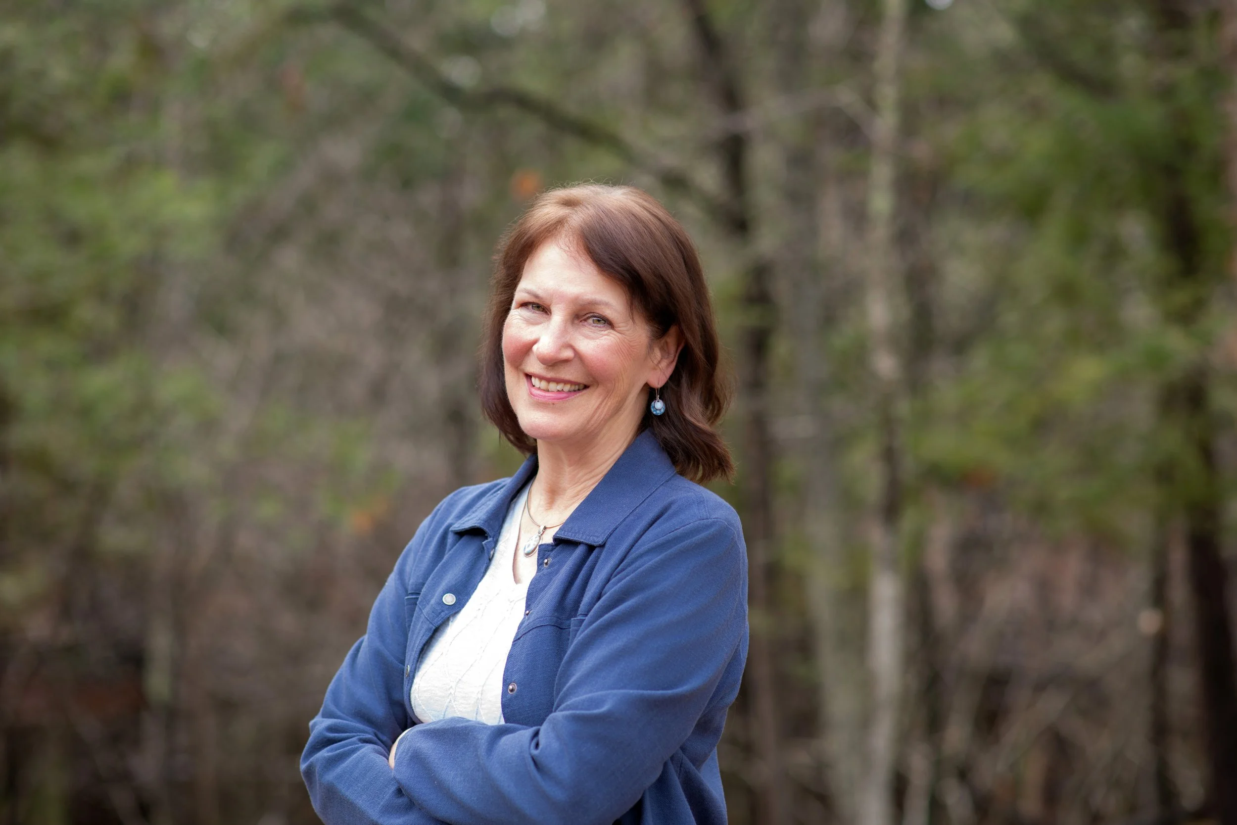 A woman with shoulder-length brown hair, wearing a blue jacket and earrings, smiling with her arms crossed outdoors in front of a blurred wooded background.