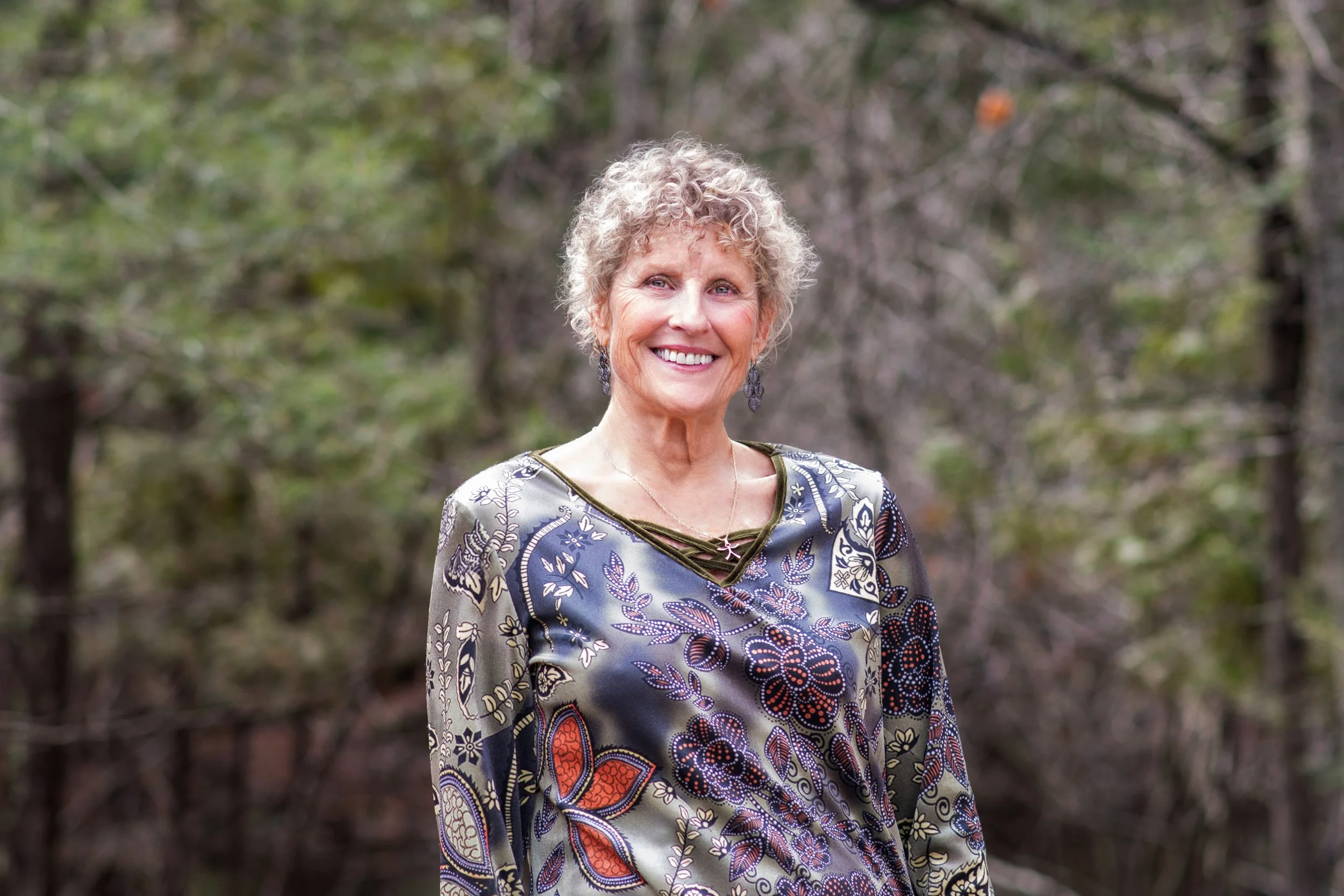A woman with short, curly gray hair smiling outdoors in front of a forested background, wearing a colorful, patterned blouse with butterfly and floral designs.
