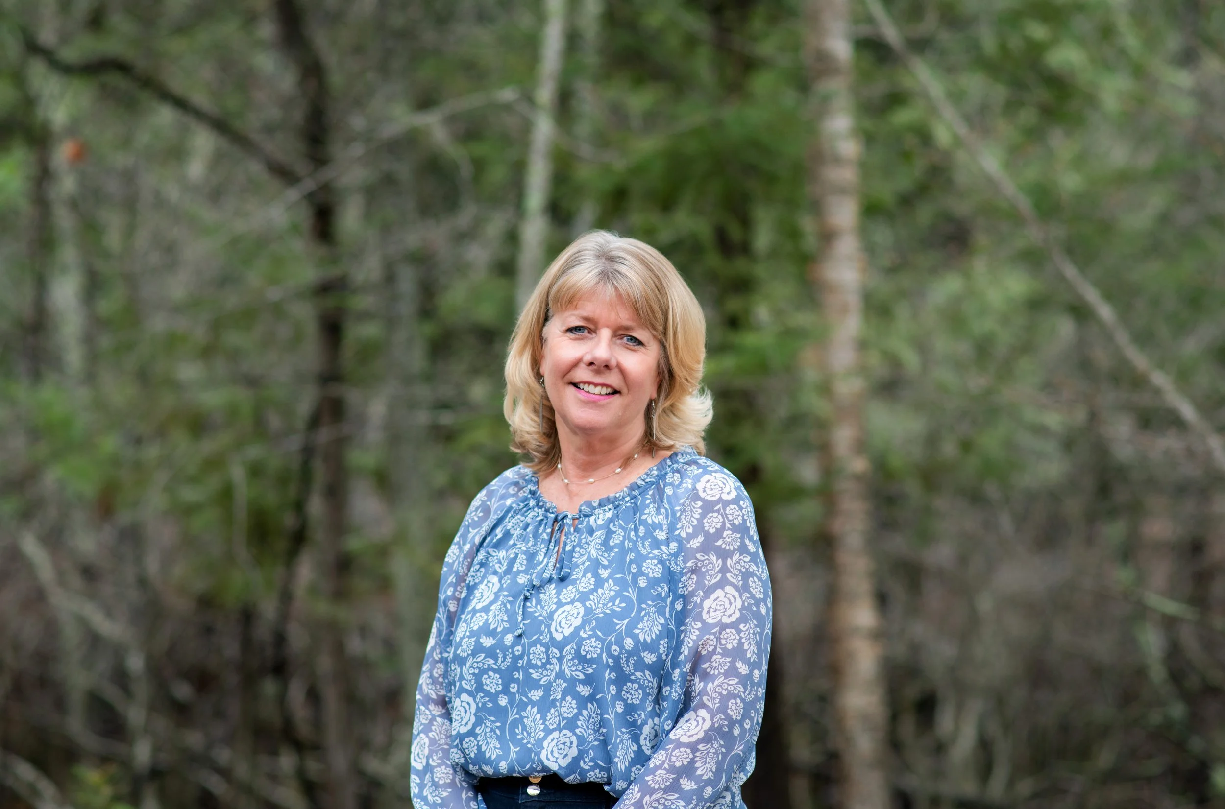 A middle-aged woman with blonde hair smiling, wearing a blue floral blouse, standing outdoors in a wooded area with green trees in the background.