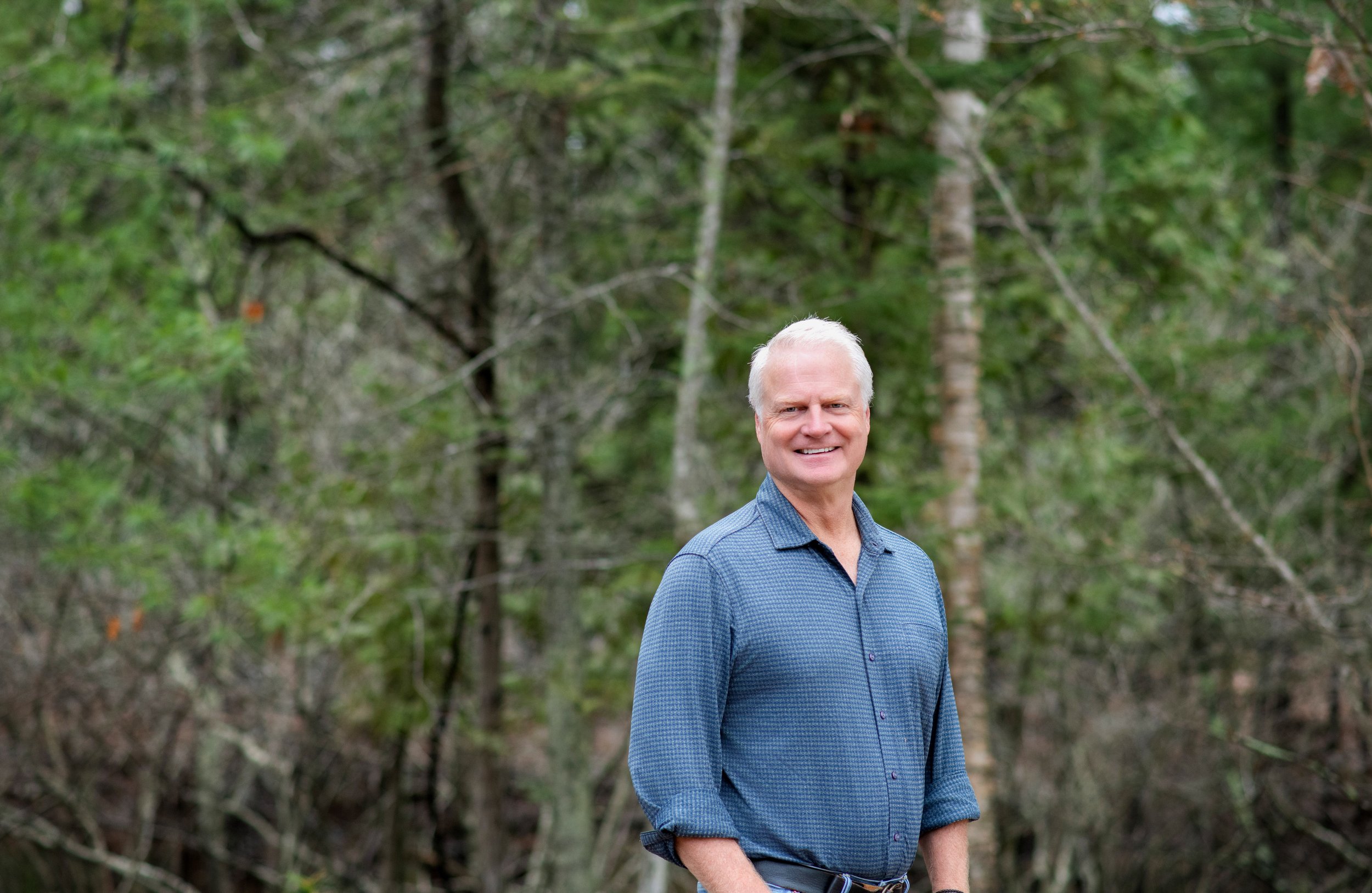 A smiling middle-aged man with gray hair wearing a blue checkered shirt standing outdoors in front of green trees.