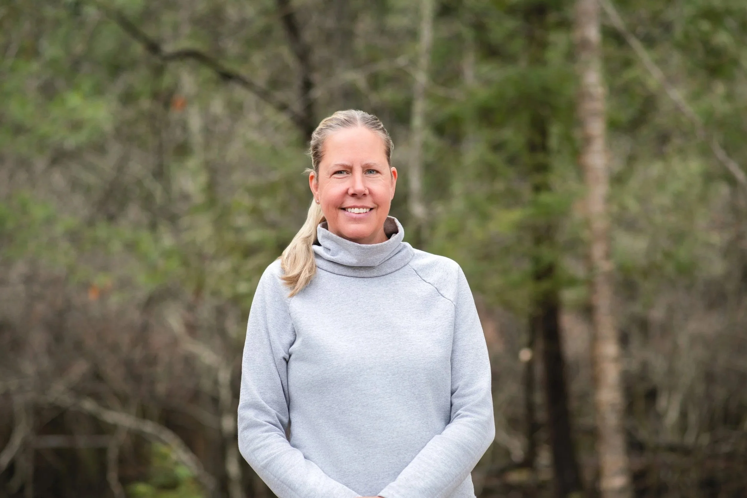 A woman with blonde hair tied back in a ponytail stands outdoors in front of a blurred background of trees and foliage, wearing a light gray turtleneck sweater and smiling at the camera.