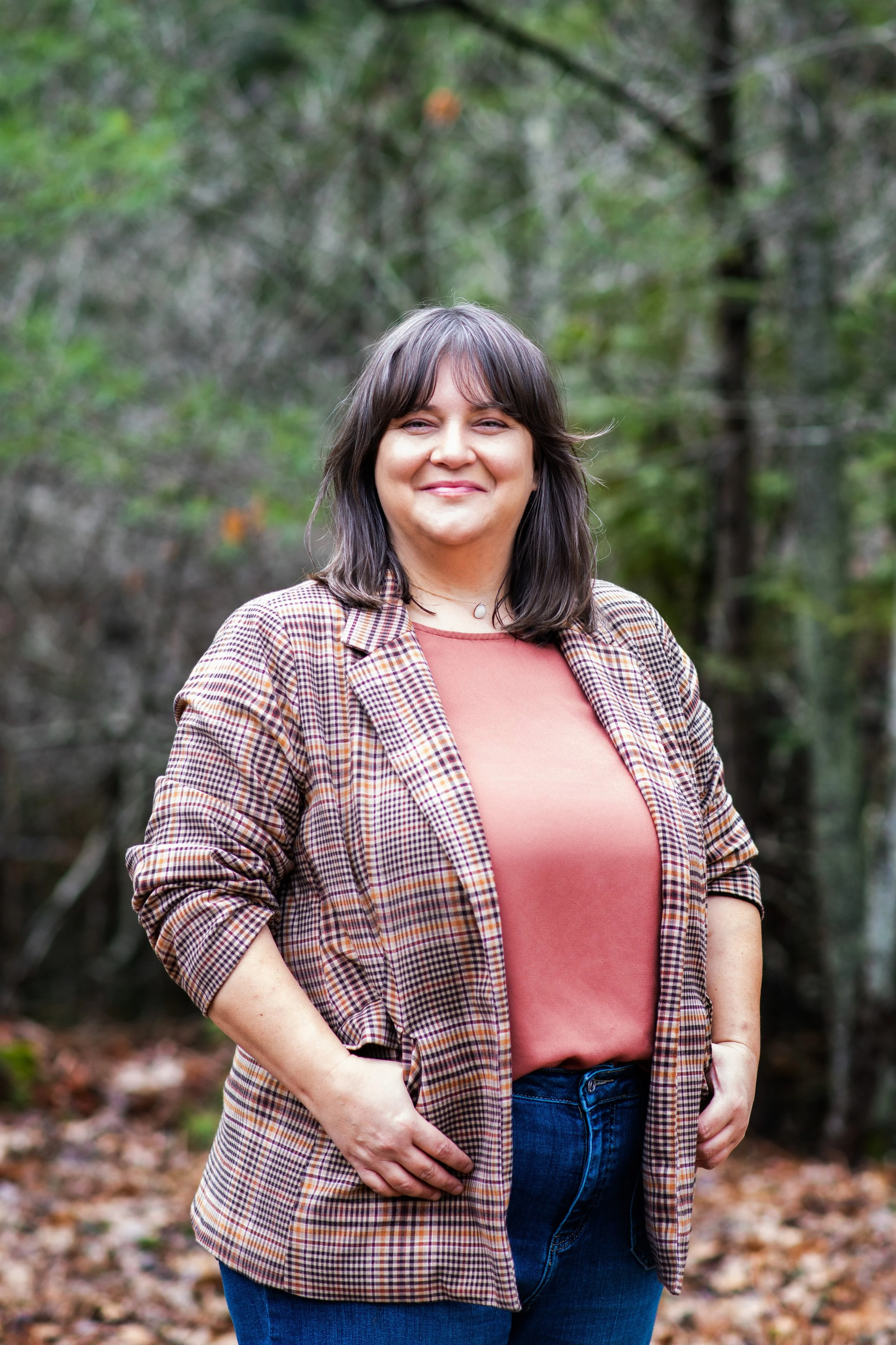 A woman with shoulder-length dark hair wearing a plaid blazer and a rust-colored top, standing outdoors in front of trees and fallen leaves, smiling.