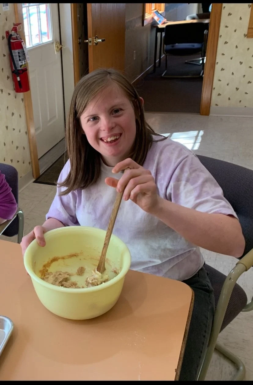 Person smiling and stirring dough in a yellow mixing bowl with a wooden spoon.