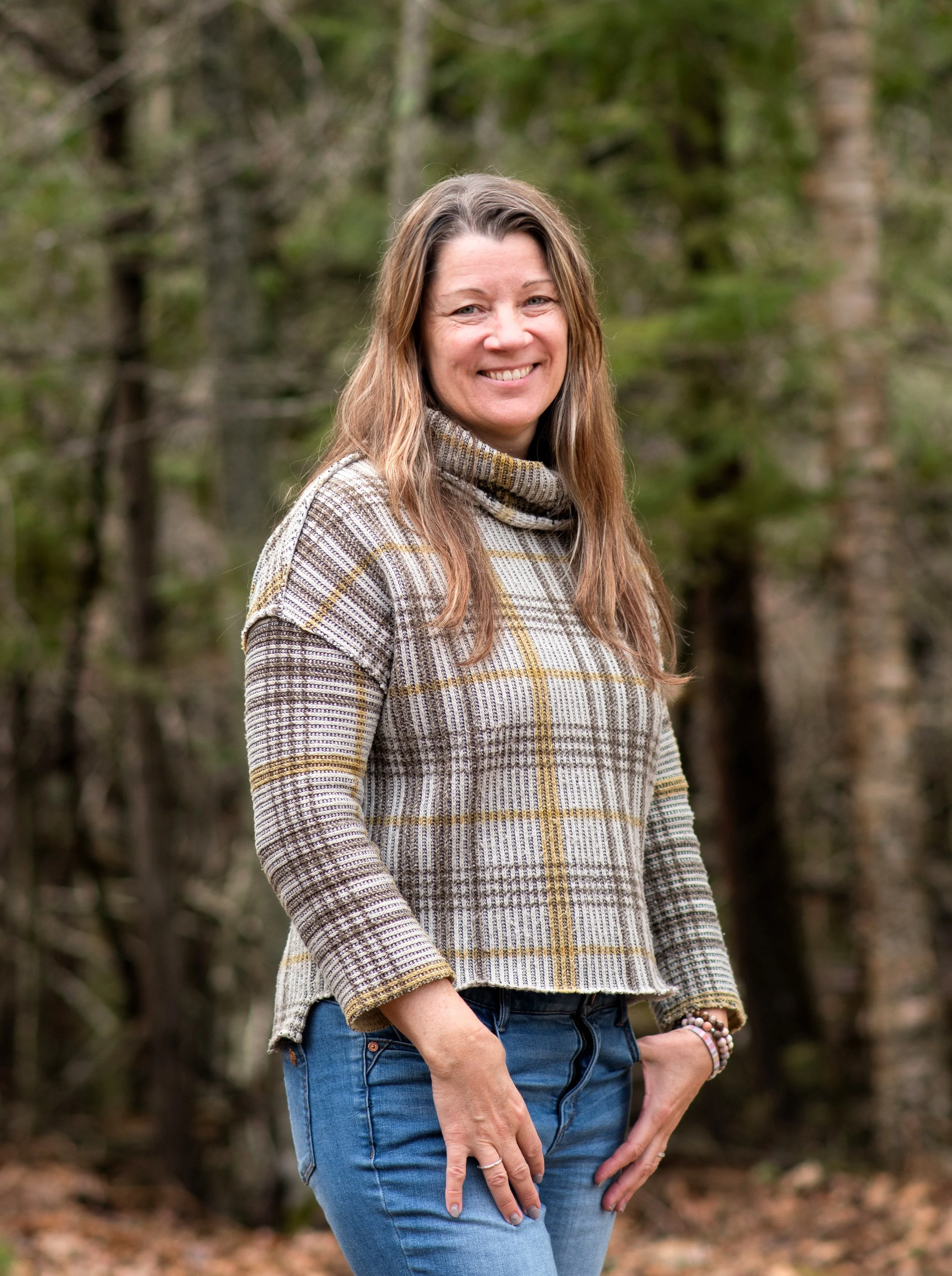 A woman with long brown hair smiling outdoors in front of trees, wearing a plaid turtleneck sweater and blue jeans.