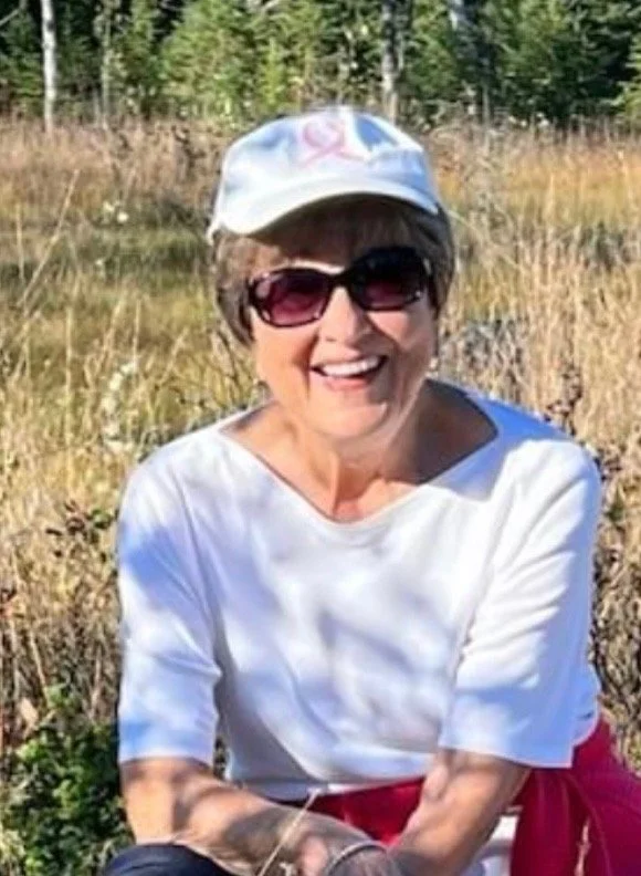 A smiling older woman wearing sunglasses, a white baseball cap, a white shirt, and a red jacket tied around her waist, sitting outdoors in a grassy area with trees in the background.
