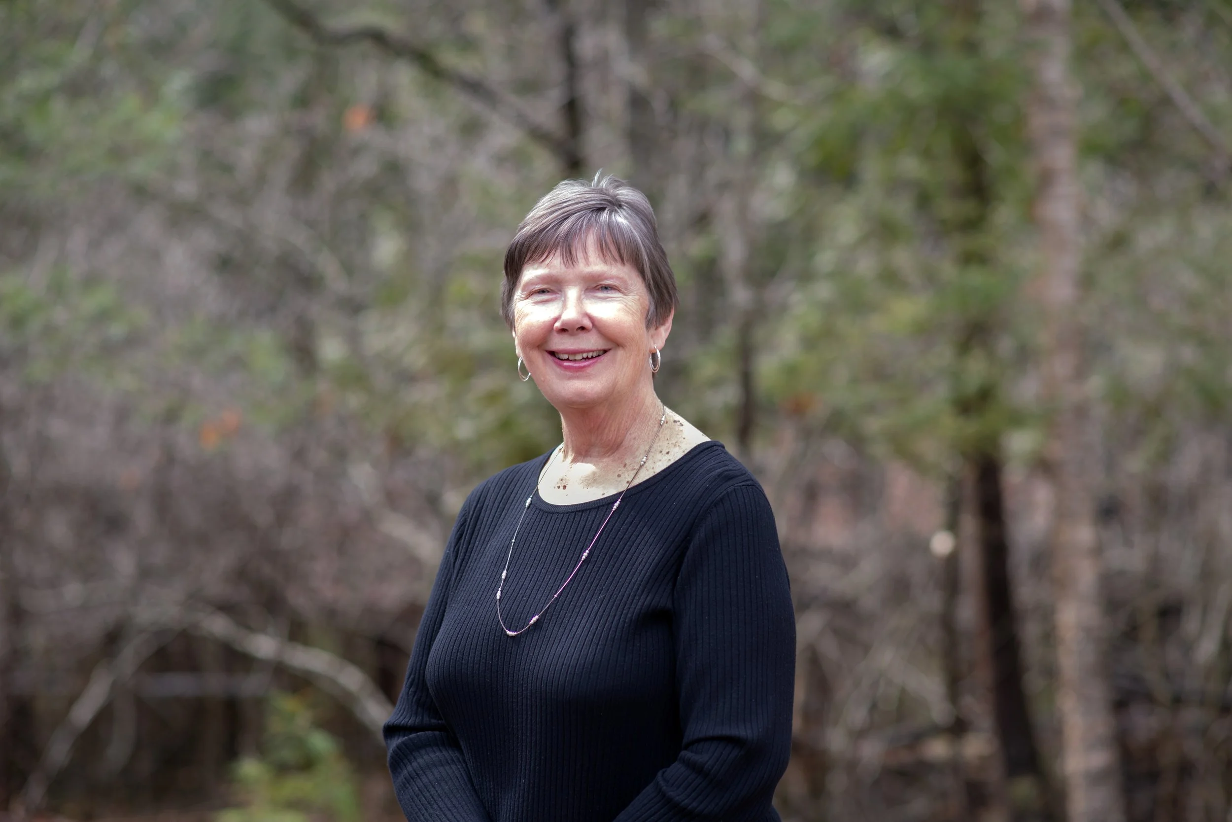 A woman with short gray hair smiling outdoors in a wooded area, wearing a black top, earrings, and a necklace.