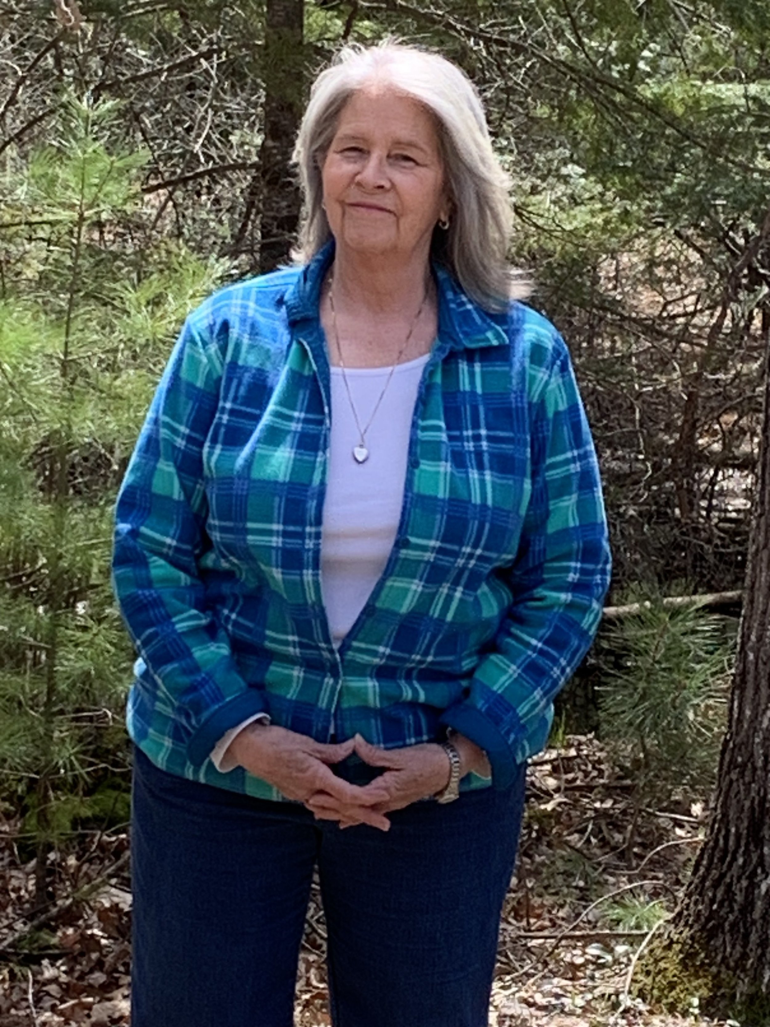 An elderly woman with gray hair, wearing a blue plaid shirt over a white top, stands outdoors in a wooded area with green trees and leaves in the background. She has her hands clasped in front of her.