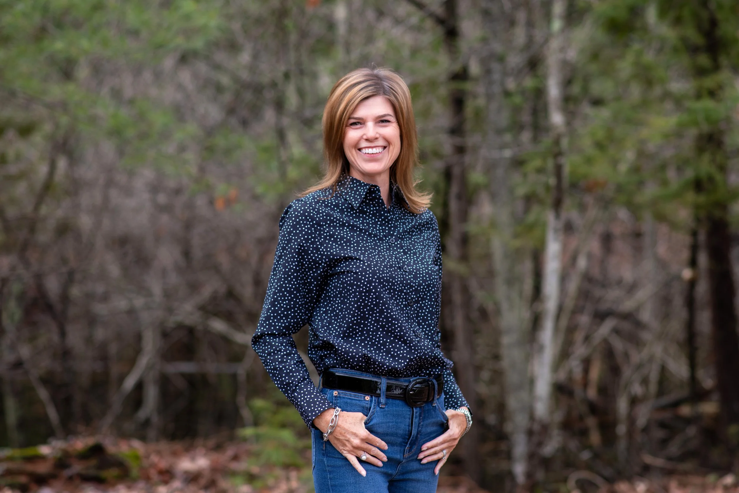 A woman with shoulder-length light brown hair smiling outdoors, wearing a dark blue shirt with white polka dots and blue jeans, standing in front of a wooded background.