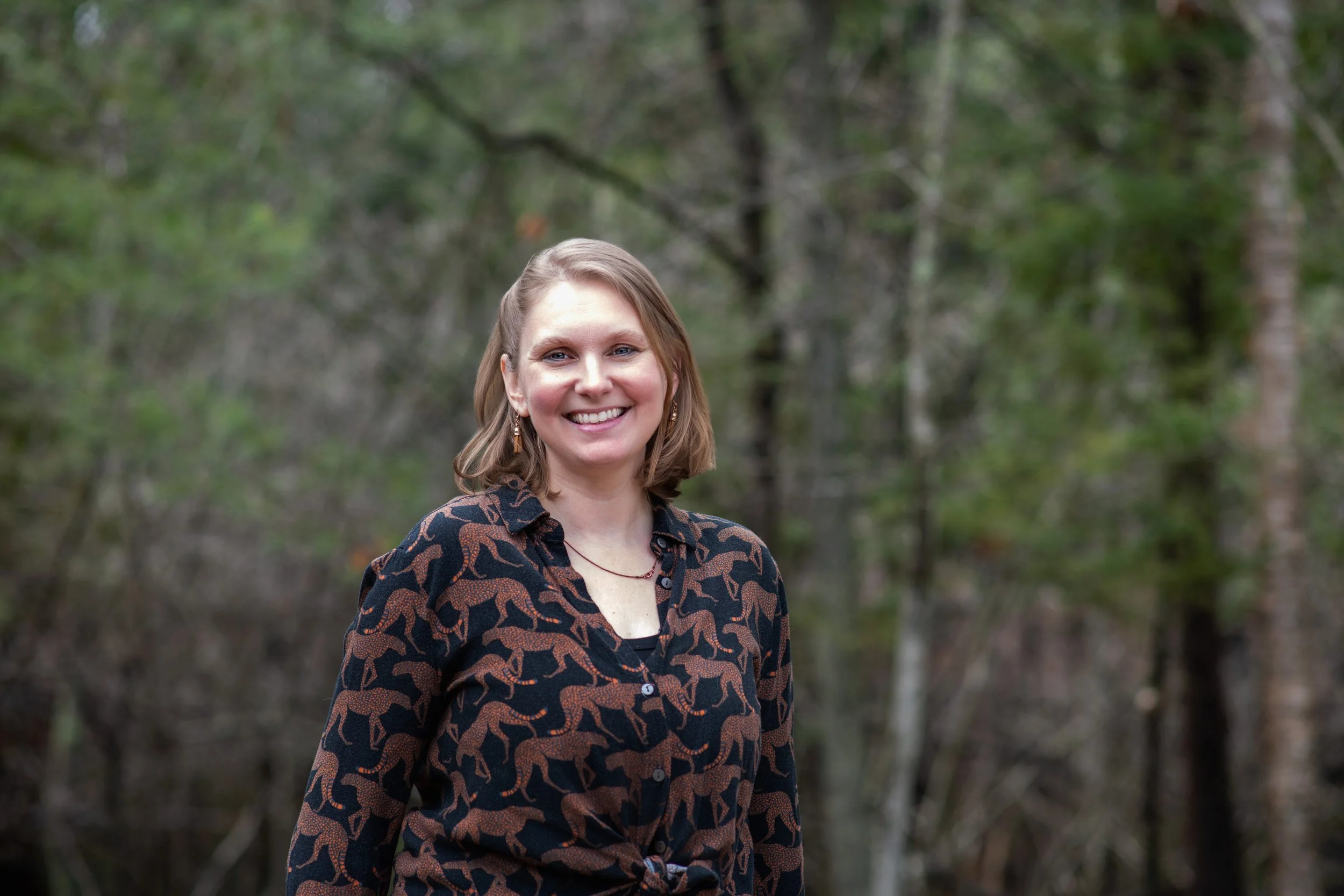 A woman smiling outdoors in a forested area with green trees in the background.