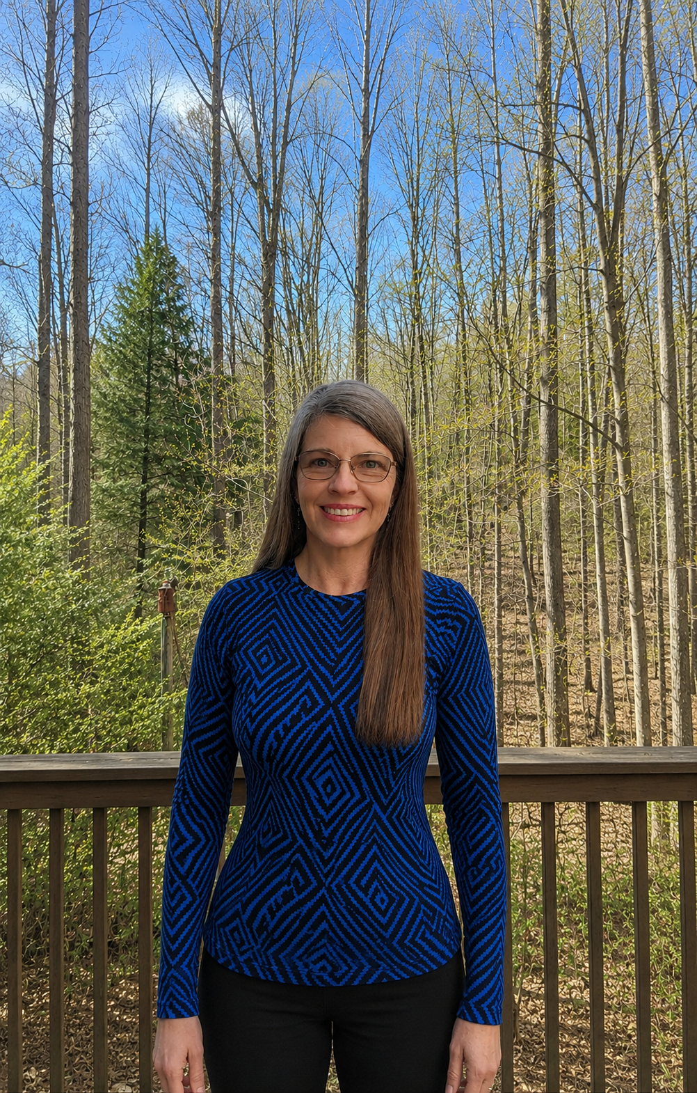 A smiling woman with glasses and long brown hair standing outside on a wooden deck, with a background of leafless trees and a clear blue sky.