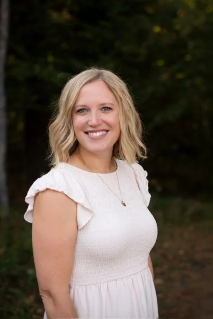 A smiling woman with blonde hair wearing a white dress standing outdoors with trees in the background.