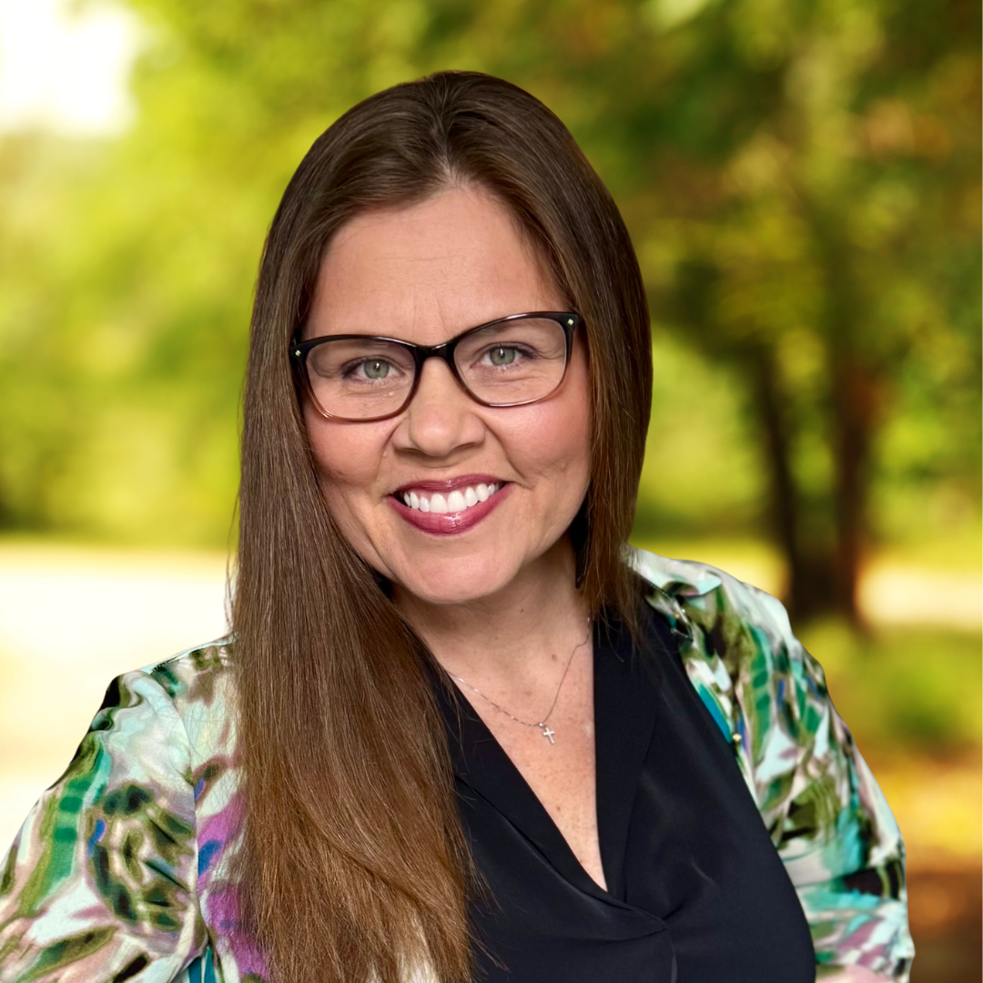 Smiling woman with glasses, long brown hair, wearing a floral-patterned blouse, in an outdoor setting with blurred greenery.